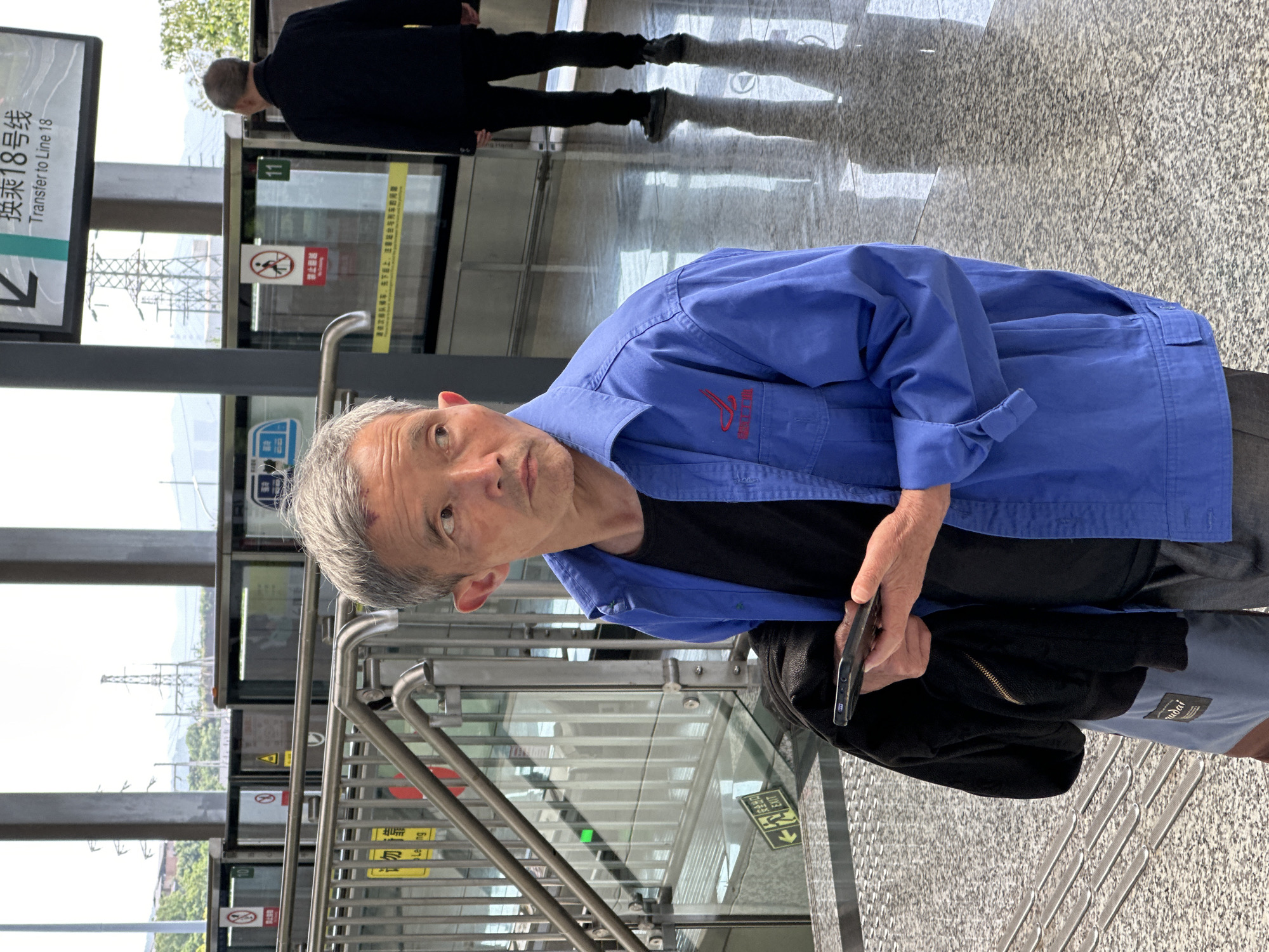 The image shows an older man in a blue jacket and black t-shirt standing on a platform, possibly at a subway station. He is holding a smartphone. Behind him, a glass barrier with automated doors displays signage in Chinese, including a sign that reads "Transfer to Line 18". There is a sign that reads EXIT. Another person in dark clothing is visible in the background, walking away. The platform has a gray, speckled floor. A window shows power lines and greenery outside. The scene is most likely taking place in Jiansheng, China, based on the language on the signs.