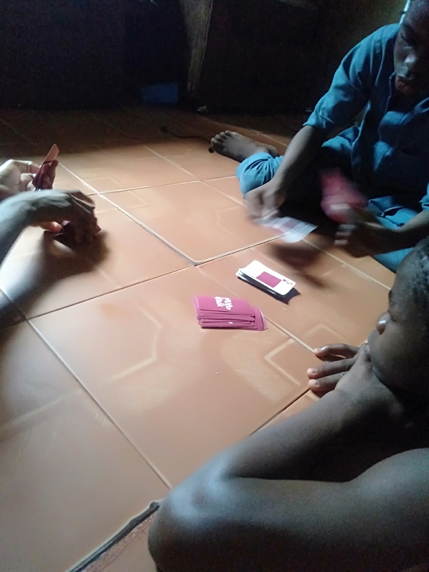 Three individuals are engaged in a card game, seated on a light brown tiled floor indoors. The scene is dimly lit, suggesting it might be evening or a poorly lit room.
On the left, a person's hands are visible, holding a fan of pink-backed playing cards. In the upper right, another person, wearing a blue shirt and trousers, sits cross-legged with bare feet, also holding cards in their hands. Their face is partially visible but blurry, showing a concentrated expression. In the lower right corner, a third person's head and arm are visible; they are looking down, with their right hand resting under their chin, observing the game.
Several stacks and individual cards are spread across the tiled floor between the players. One prominent stack of cards features pink/magenta backs. Among the visible cards, the game "UNO" is identifiable, with some cards clearly displaying the "UNO" logo, numbers like "5", and action symbols such as "SKIP". The cards on the floor include both pink-backed cards and some white-faced cards.
The setting appears to be a casual, relaxed environment, likely a home in Shangisha, Nigeria, where the participants are focused on the card game. The dark background behind the players indicates the depths of the room.