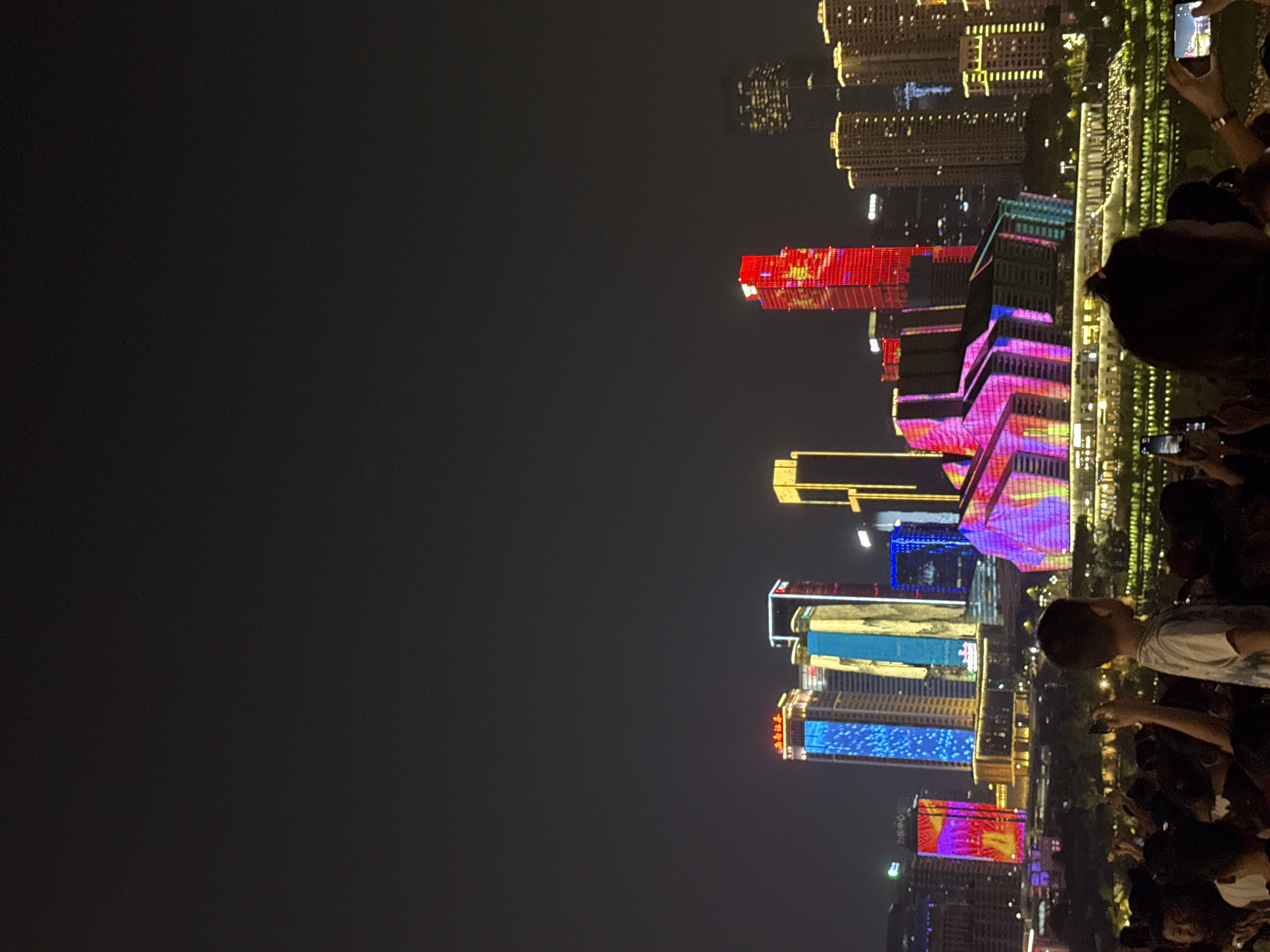 An elevated, eye-level, medium shot captures a night cityscape in Yuzhong District, China, featuring a vibrant display of illuminated skyscrapers and buildings. In the foreground, a crowd of people are gathered, some holding up smartphones to capture the scene. Among them, a young boy stands out, his back to the camera, wearing a tie-dye t-shirt. The background is dominated by the architecture of the city, with buildings adorned in various colors, including bright blues, yellows, pinks, and reds. One prominent red skyscraper stands out, with patterns projected onto its facade. In the distance, a cluster of taller, lighter-colored buildings are visible, also illuminated. The overall impression is one of a lively urban environment at night, possibly during an event or celebration.