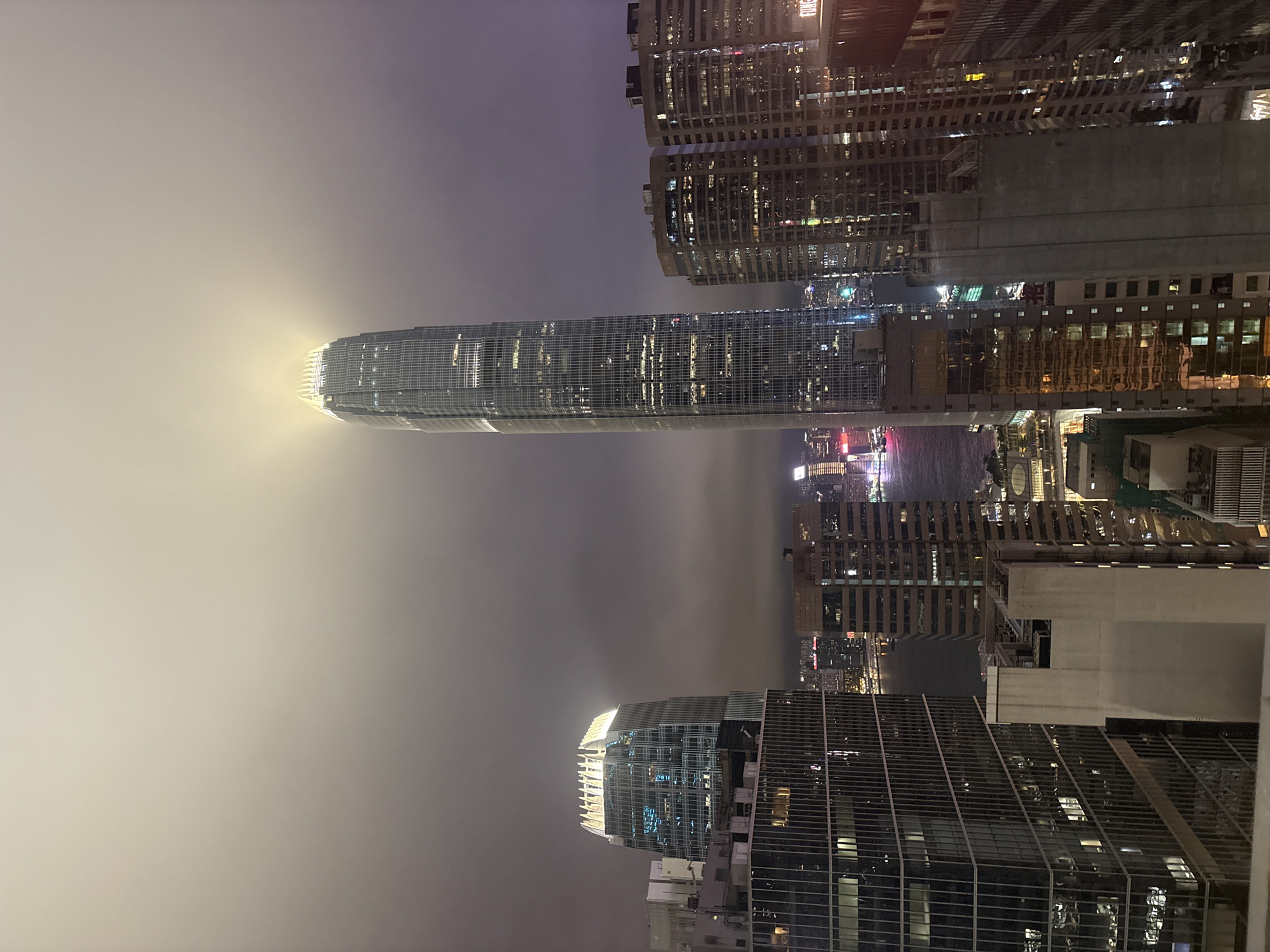 This is a nighttime view of Hong Kong's skyline shrouded in heavy fog or smog. The image appears to have been taken from an elevated position, possibly a high-rise building. Several skyscrapers are visible, with the most prominent one appearing to be the International Finance Centre (IFC), recognizable by its distinctive shape and the illuminated top section. The buildings are densely packed, reflecting the urban density of Hong Kong. Some light trails suggest traffic activity, even though obscured by fog. The sky is dark and indistinct due to the heavy fog.