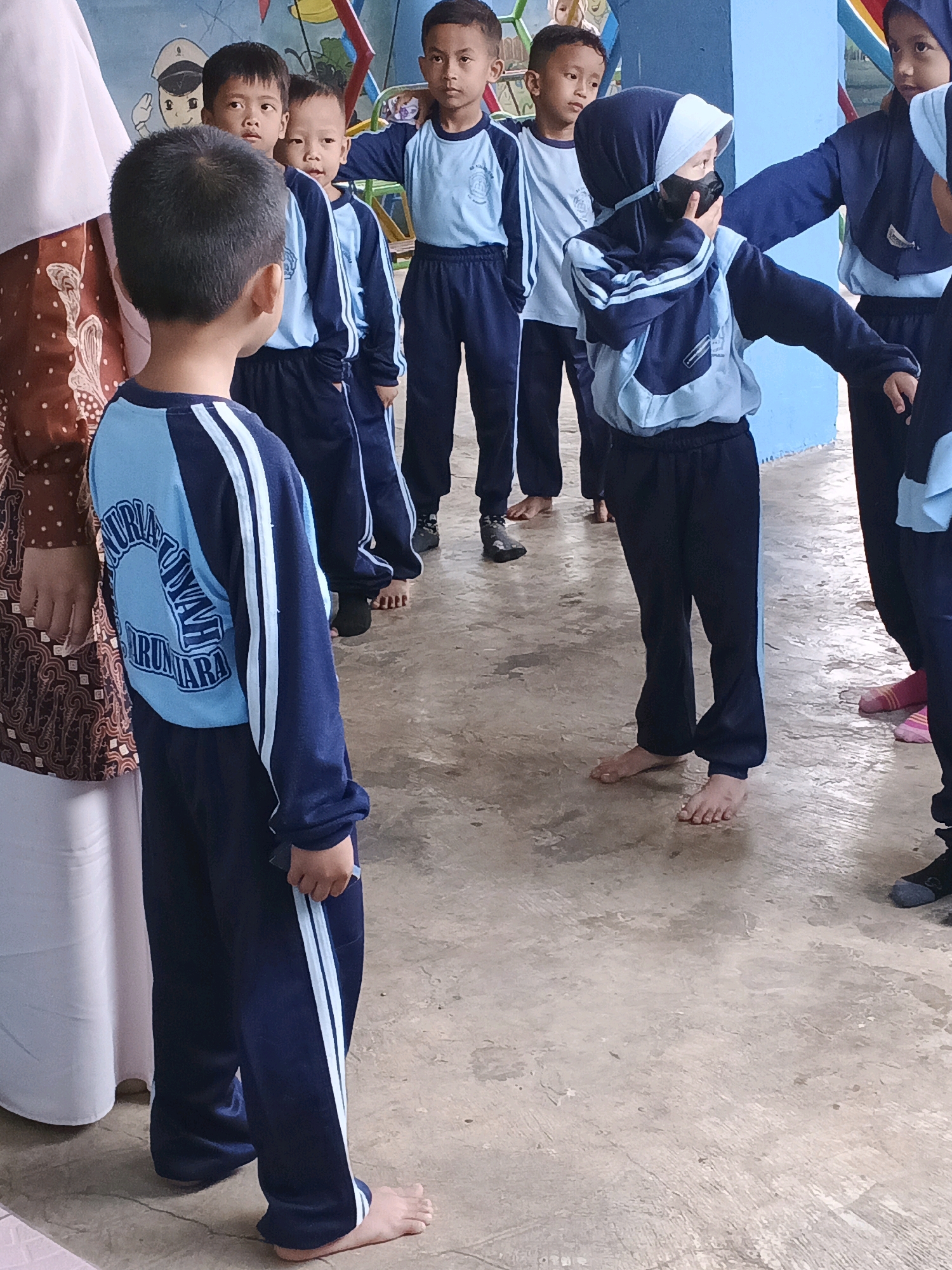 The image depicts a group of children, likely students, standing in a line outdoors. They are all wearing matching navy blue and light blue uniforms. The child in the foreground on the left is facing away from the camera, his back to the viewer, and is wearing a long-sleeved uniform shirt with a circular logo on the back that appears to read "TURIYOKUL SUMBARA". The other children are facing towards the camera or to the side. One child, wearing a hijab and a face mask, is looking to the side and touching their mask. Some of the children are barefoot, while others are wearing shoes or socks. The background shows a colorful mural on a wall, suggesting the location might be a school or playground. The ground is concrete and appears wet in places, indicating it may have rained. Based on the visible text on the uniform, the location is identified as Warungkiara, Indonesia.