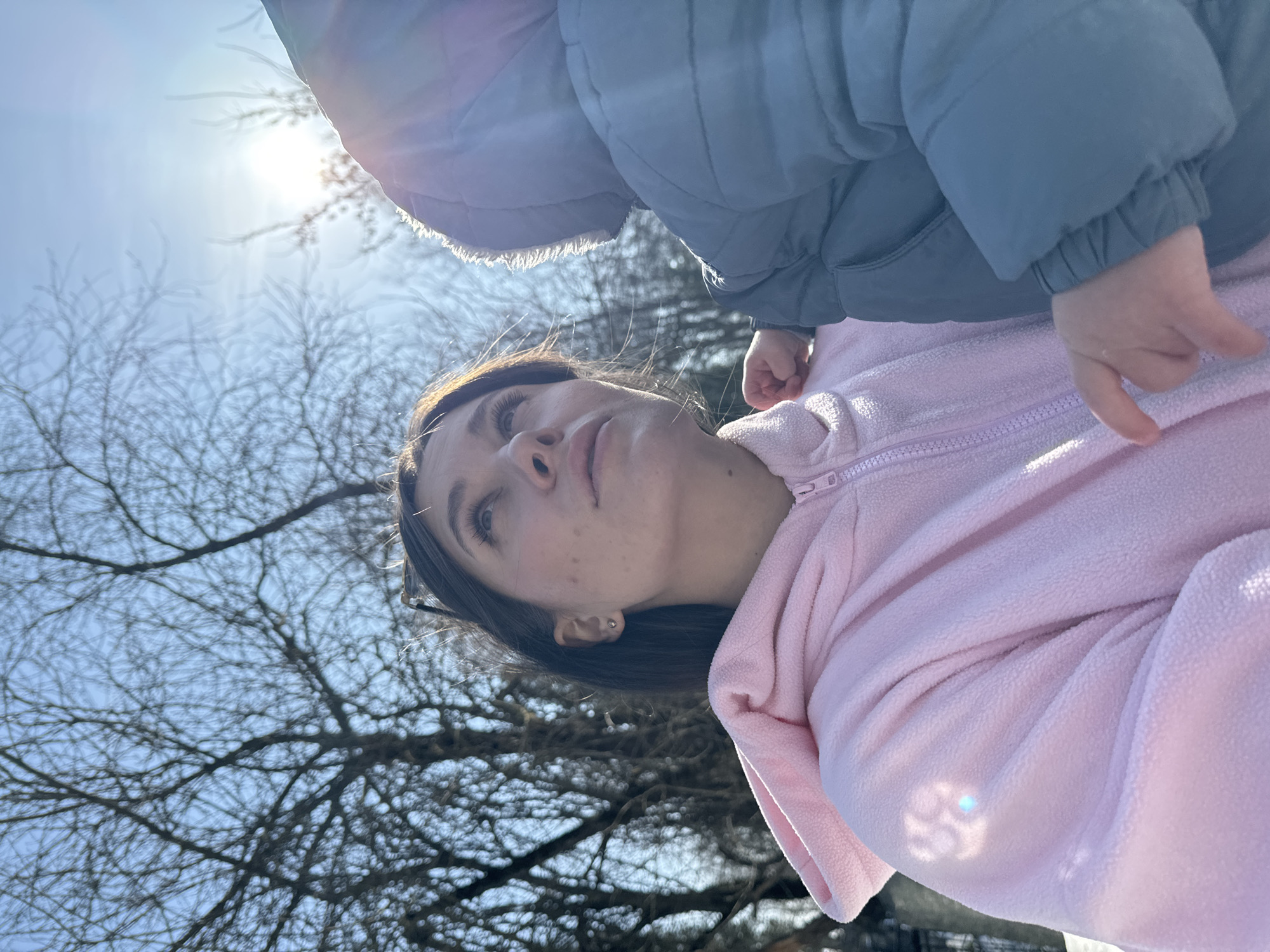 The image depicts an outdoor scene, reported to be in Lovtsovo, Russia, on a bright, sunny day. An adult female is positioned in the left and central foreground, looking upwards and to the right. She has dark hair, a light complexion with visible freckles, and wears a pink, full-zip fleece hoodie. A small, round stud earring is visible in her left ear, and a pair of sunglasses rests on her head. To her right, a young child is partially visible from the back, wearing a grey or dark blue padded winter jacket with a hood featuring a fuzzy trim. The child's right hand is resting on the woman's left shoulder/chest area. The background features a clear blue sky and numerous bare deciduous trees. The sun is prominently visible in the upper right quadrant, emitting strong glare and light across the scene. White areas at the bottom of the frame suggest ground cover, possibly snow.
