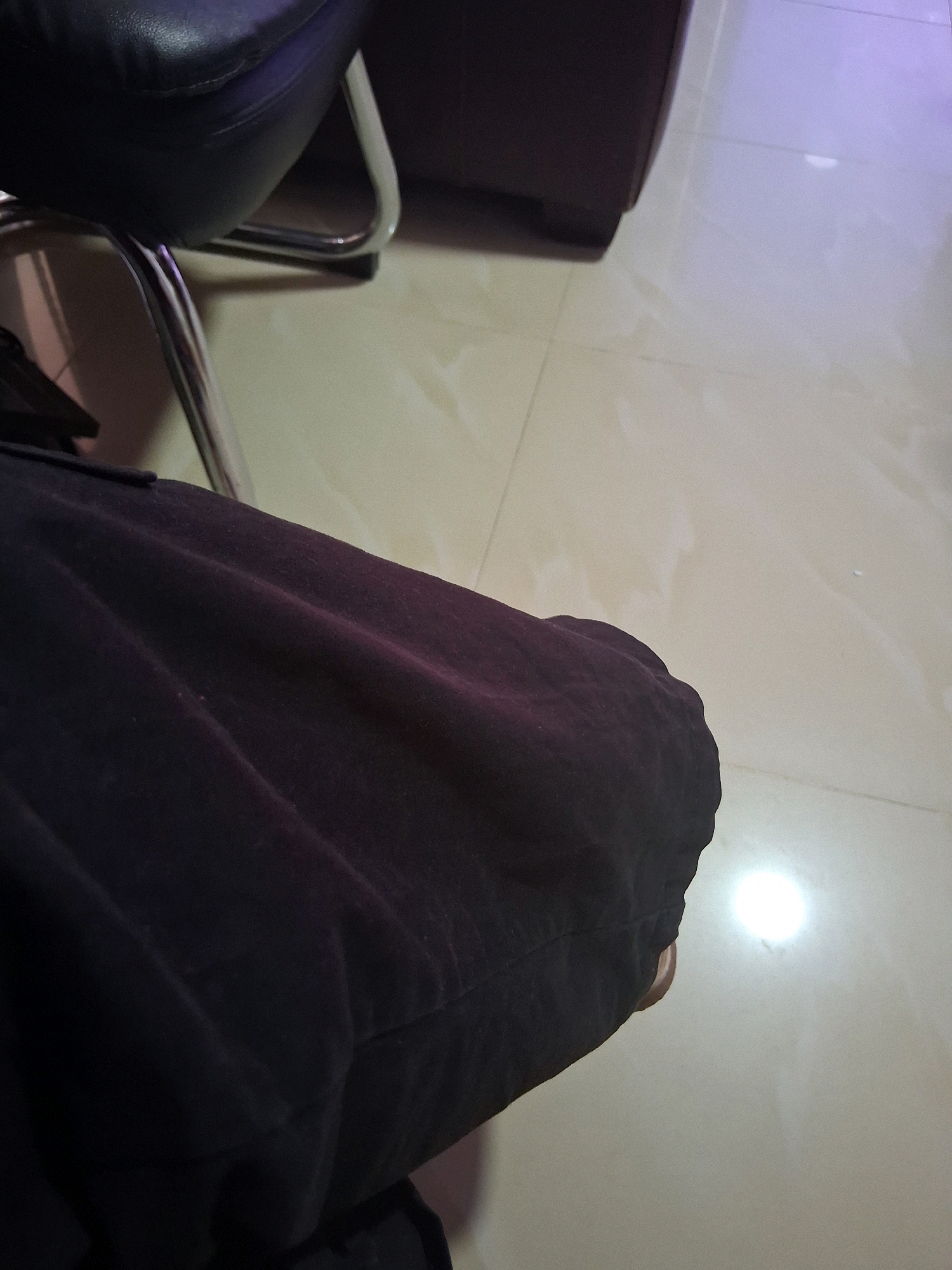 The image shows a close-up view of a person's leg wearing dark pants, resting on a metallic chair in what appears to be an indoor setting. The floor is tiled with a light-colored, reflective surface, possibly marble or porcelain, common in public spaces or homes in La Vega, Dominican Republic. The lighting suggests artificial illumination, with a bright reflection on the floor. The background includes a glimpse of the chair's metallic frame and a dark, possibly leather, seat. No specific landmarks or street views are visible to pinpoint the exact location within La Vega, but the overall environment suggests a residential or commercial interior.
