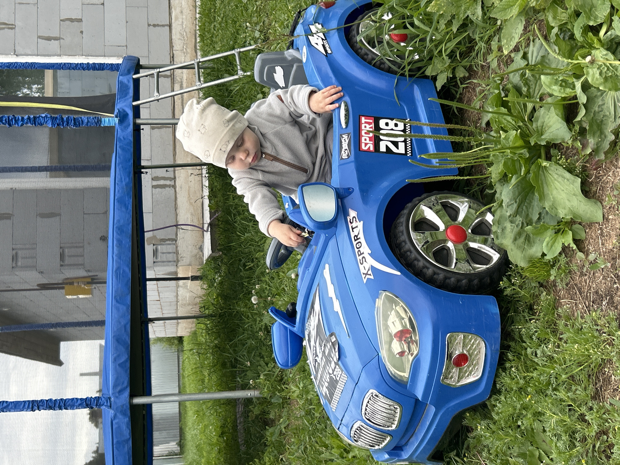 A young child wearing a light-colored beanie and a grey sweatshirt is sitting in a bright blue toy car. The car has "SPORT 218" written on its side, along with other racing-themed decals. The child's hands are on the steering wheel. The car is parked on a grassy area with some plants growing around it. In the background, there is a building under construction, with white brick blocks and scaffolding. A blue and black trampoline net is also partially visible to the left of the image, with metal poles supporting it. The scene appears to be outdoors, likely in a backyard or a yard adjacent to a construction site. The lighting suggests it is daytime. The child's gaze is directed downwards, and their expression is difficult to determine due to the angle and their closed eyes.