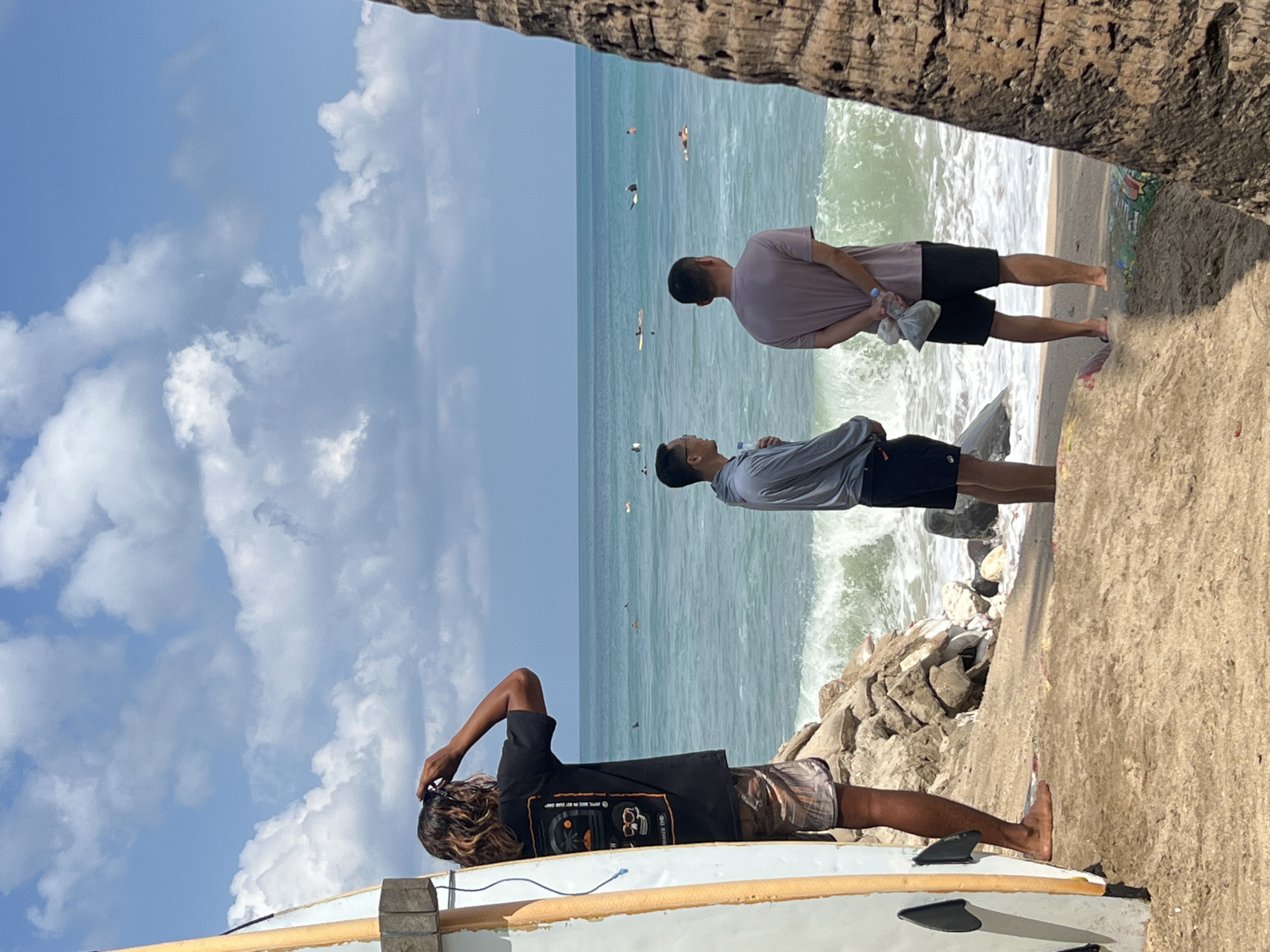 The image shows a beach in Kuta, Indonesia, with three men and a surfboard. A man on the left is wearing a black t-shirt and patterned shorts, with a surfboard standing next to him. He is looking out at the ocean and holding sunglasses up to his eyes. Two other men are standing in the center of the image, facing the ocean. The man on the left is wearing a grey long-sleeved shirt and black shorts, holding a bottle of water. The man on the right is wearing a purple t-shirt and black shorts, holding two plastic bags. The ocean is visible in the background, with several surfers in the water. The sky is blue with scattered white clouds. The foreground consists of sand and some rocks.
