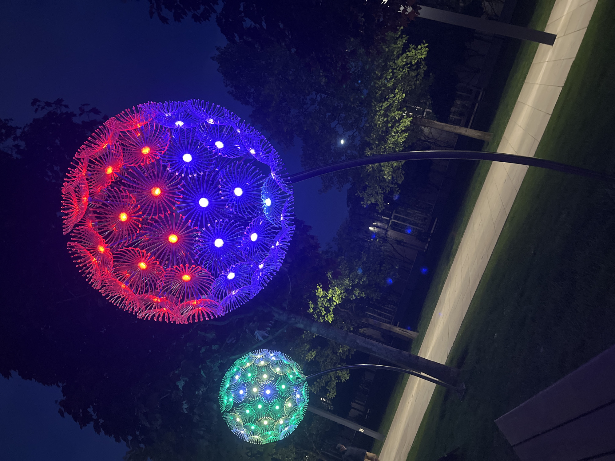 The image shows two large, spherical, decorative light installations in an outdoor setting. The top sphere is illuminated with red and blue lights, while the bottom sphere is illuminated with green lights. They are mounted on arched metal poles. The surrounding environment includes trees, grass, a sidewalk, and what appears to be a bench. The scene is illuminated by the light installations and possibly other ambient light sources. The location is Krasnodar, Russia.
