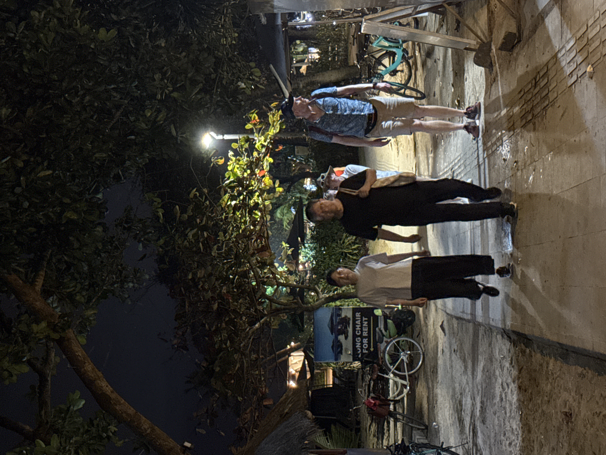 The image depicts a nighttime scene on a paved walkway adjacent to a sandy area, likely a beachside path in Denpasar, Indonesia. Several individuals are walking along the path, illuminated by streetlights and ambient lighting from nearby establishments. On the left side of the walkway, there is a sign that reads "LONG CHAIR FOR RENT" with an image of lounge chairs on a beach. Various bicycles and possibly a scooter are parked in the background, some with red bags or baskets attached. Trees with lush foliage overhang the walkway, creating a dappled lighting effect. In the distance, there appear to be structures with thatched roofs and lights, suggesting a commercial or recreational area. The people are casually dressed, and the overall atmosphere is one of evening activity in a tropical setting.