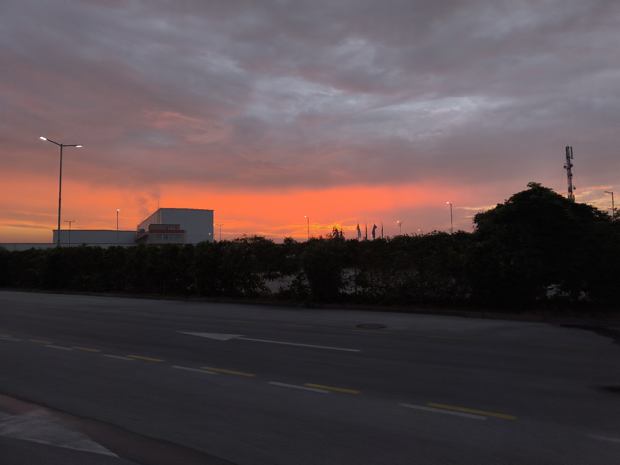 An outdoor scene captures a vivid sunset or sunrise over what appears to be an industrial or commercial area in Benkovski, Bulgaria. The sky is the dominant feature, displaying a spectacular gradient of intense orange and red near the horizon, fading upwards into softer pinks, purples, and grey clouds.
In the middle ground, several artificial lights are illuminated, suggesting dusk or dawn. To the left, a prominent streetlight stands tall. Behind a dark line of dense hedges or trees, which runs parallel to the road, a large, rectangular white building with reddish-brown elements is visible. A faint wisp of smoke or steam appears to emanate from its roof. Further to the right, partially obscured by trees, are more buildings and several tall poles flying indistinguishable flags. On the far right, a tall, dark communication tower rises above a cluster of dark trees, silhouetted against the colorful sky.
The foreground features a wide, asphalt road with clear markings. Dashed white lines and parallel dashed yellow lines segment the road, and a white arrow painted on the surface indicates a forward direction. A manhole cover is visible on the right side of the road. There are no vehicles or people present, contributing to a sense of quietude. No discernible text is visible anywhere in the image. The scene presents a dramatic interplay between the natural beauty of the sky and man-made infrastructure.
