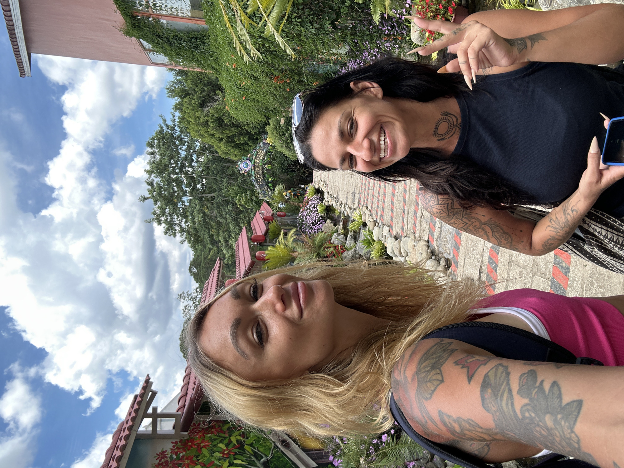 Two women are taking a selfie in front of a "Fairytale Land" attraction in Da Lat, Vietnam. The woman on the left has blonde hair and is wearing a pink tank top. She has tattoos on her arms and shoulder. The woman on the right has dark hair and is wearing a black tank top. She has tattoos on her arms and neck, and is holding up two fingers in a peace sign. The background shows a pathway lined with flowers and greenery, and buildings with red roofs. The sky is blue with white clouds.