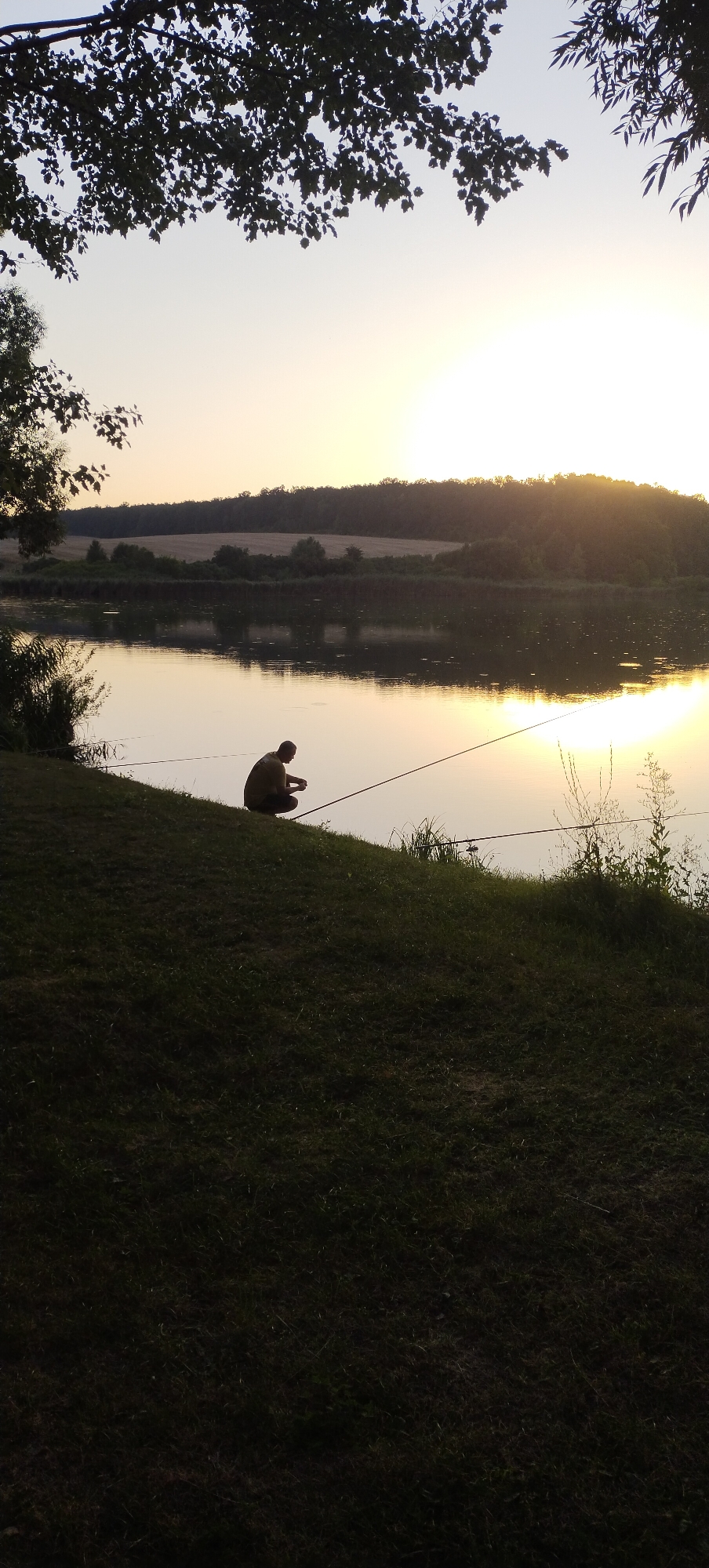 A lone fisherman is seen crouching on the grassy bank of a calm river at dusk. The setting sun casts a warm glow on the water, creating a reflective surface. The distant shoreline is lined with trees and rolling hills, under a pale sky. The foreground is dominated by a dark, grassy slope, with a few weeds growing near the water's edge. The fisherman is focused on his activity, with fishing lines extending into the water. The scene is tranquil and evokes a sense of peaceful solitude. The location is identified as Vysokopillia, Ukraine.