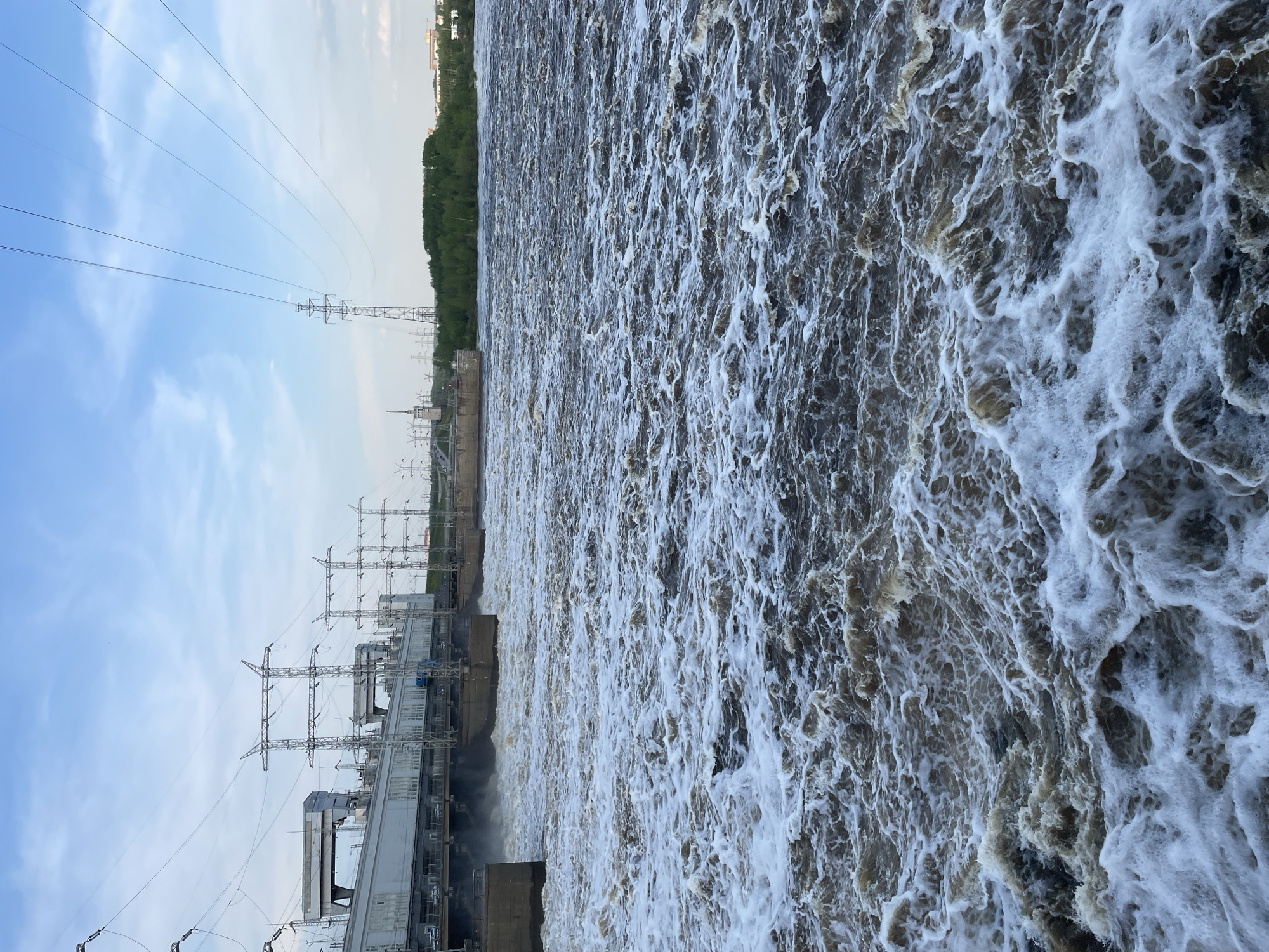 The image depicts a hydroelectric dam in Perm, Russia, with a massive outflow of turbulent, frothy water. Power lines and electrical infrastructure are visible on the dam's left side. Trees line the river bank above the dam. The sky is partly cloudy.