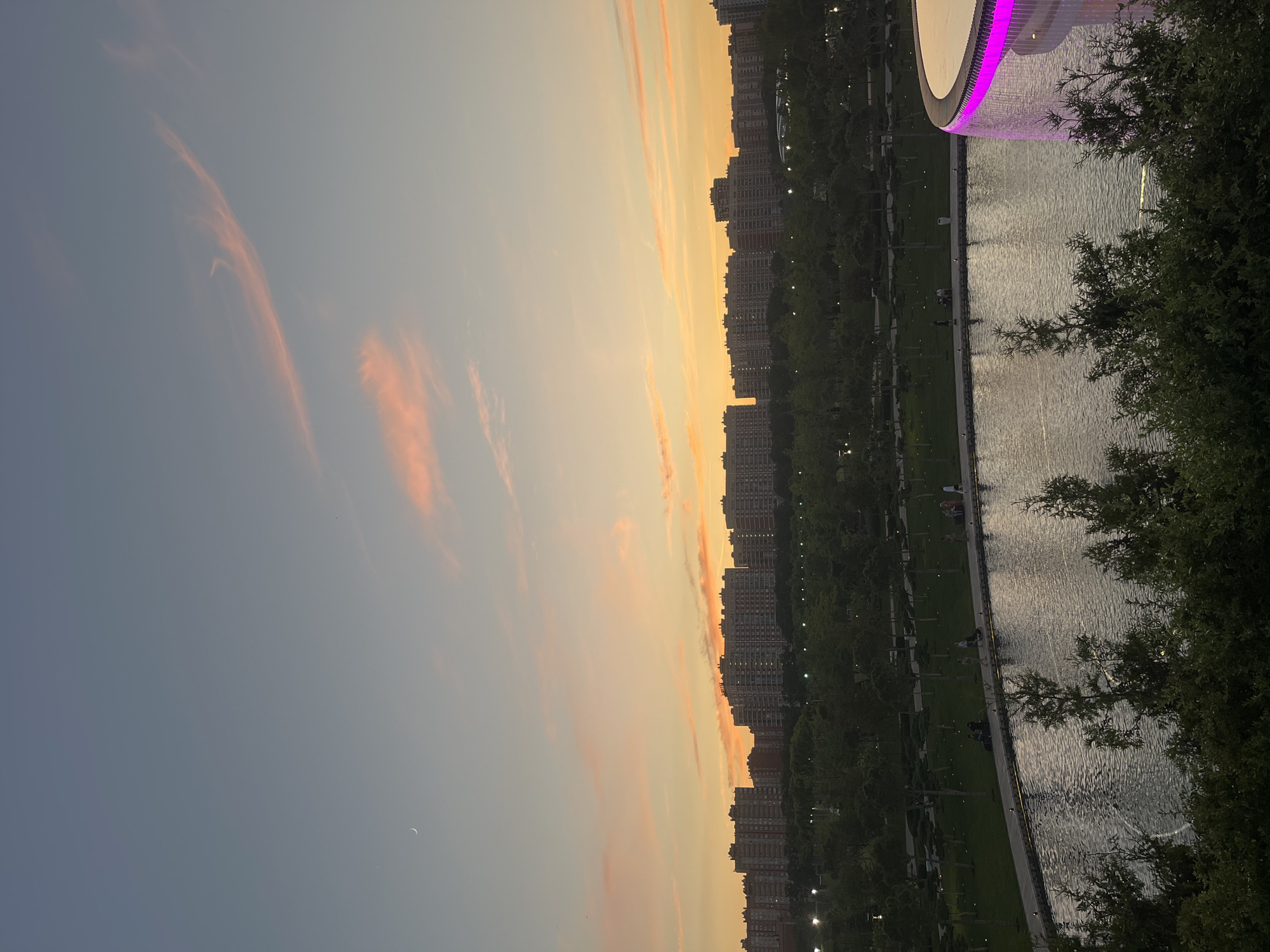 The image captures a sunset over a city park in Krasnodar, Russia. In the foreground, lush green trees partially obscure a tranquil body of water. The water's surface reflects the soft hues of the twilight sky, which transitions from pale blue at the top to a warm orange and pink gradient towards the horizon. A crescent moon is visible in the upper left quadrant of the sky.
In the midground, a paved pathway curves along the edge of the water, with a few silhouetted figures strolling or sitting along its length. Beyond the pathway lies a manicured park with neatly trimmed grass and rows of trees, illuminated by subtle lighting fixtures. In the background, a dense line of apartment buildings stretches across the horizon, their windows reflecting the fading daylight. To the far right, a modern structure with vibrant magenta lighting encircles its rim, likely part of a water feature or architectural element. The overall scene evokes a peaceful evening ambiance in an urban park setting.