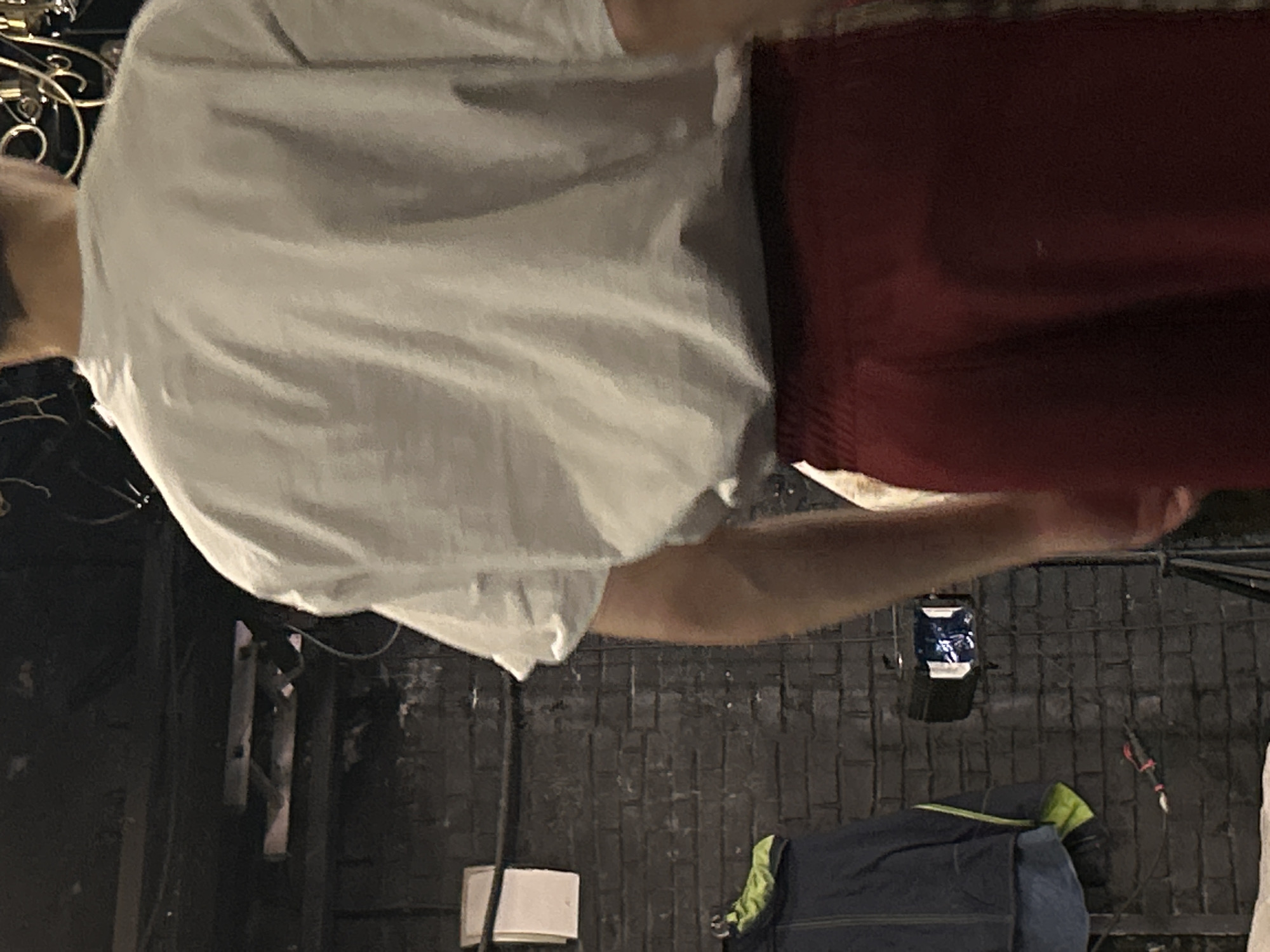 A person, seen from the back, stands in a dimly lit room. They are wearing a light-colored t-shirt and dark red pants. The background features a dark brick wall with some lighting equipment, including a spotlight and a hanging lamp. A jacket and a piece of paper are also visible on the wall.
