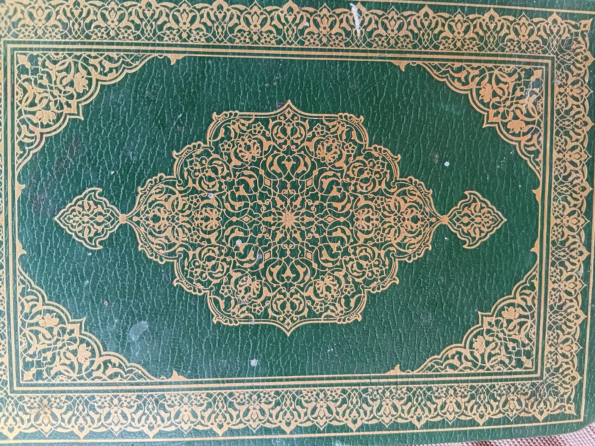 The image displays a close-up view of a book cover, showcasing intricate gold ornamentation on a dark green background. The cover is rectangular and features a prominent border made of repeating gold arabesque or floral patterns. Inside this broad decorative border, a thin gold line further frames the central area.
At the center of the cover is a large, elaborate, and symmetrical gold design. This central motif is highly detailed, composed of interconnected arabesque and possibly geometric patterns, forming an elongated, medallion-like shape with pointed extensions on the horizontal axis. Smaller, complementary gold decorative elements extend outwards from the sides of the central design, enhancing the overall ornate composition.
The dark green surface of the cover has a distinct pebbled or grained texture, possibly indicating a material like leather or a textured binding cloth. There are a few minor scuffs and small white specks visible on the green background, suggesting some age or use of the book. No text is visible anywhere on the cover. The style of the ornamentation is characteristic of traditional Islamic art and bookbinding, aligning with the cultural context of Martil, Morocco. The image captures the elegance and detailed craftsmanship of the cover design.