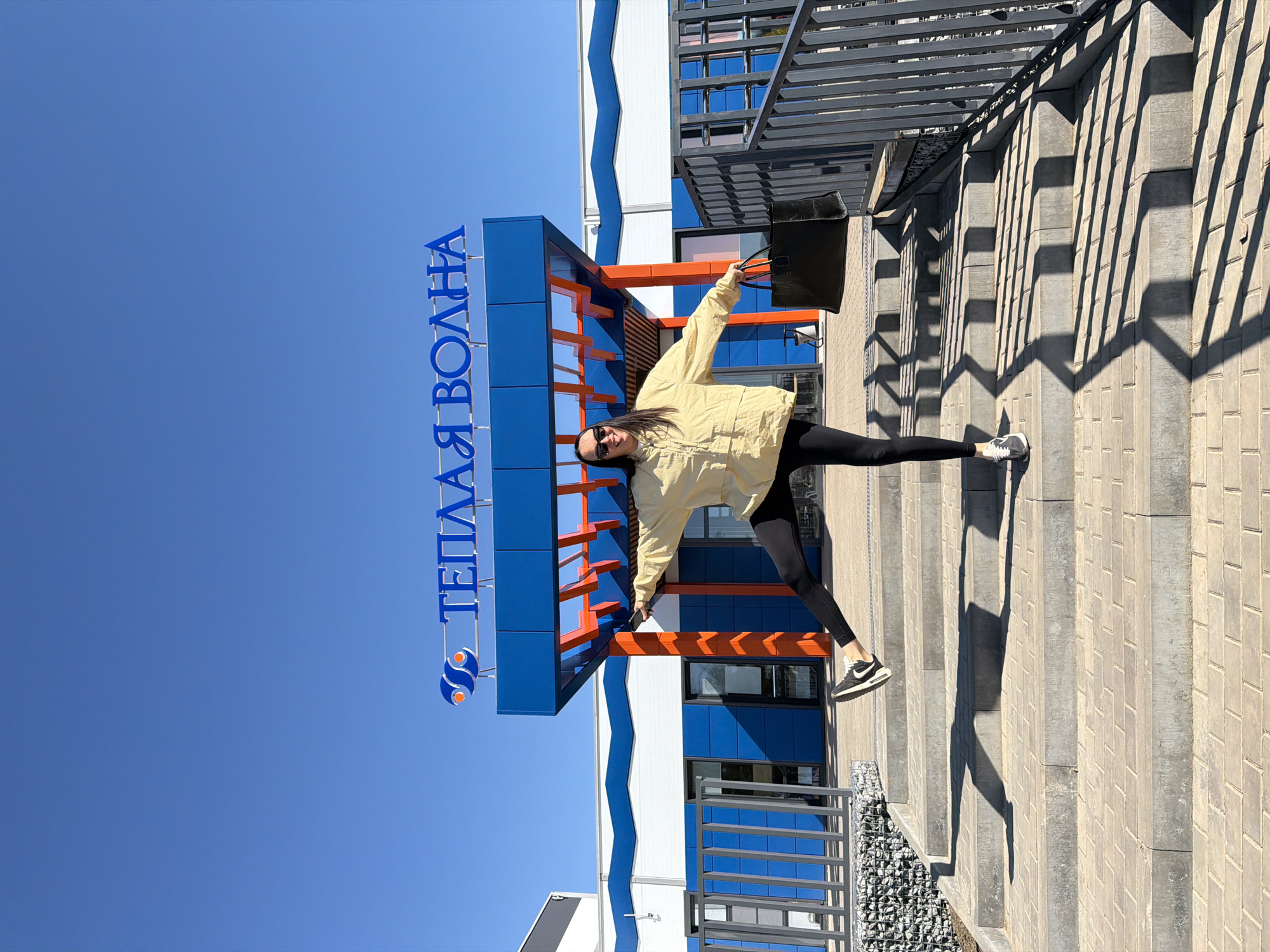 A medium shot of a woman standing on a flight of stairs in front of a building with the inscription "ТЕПЛАЯ ВОЛНА", located in Votkinsk, Russia. The woman is wearing black leggings, a light yellow jacket, and black and white sneakers. She is holding a black bag. The building is primarily blue and white with orange accents. The sky is clear and blue. There are shadows cast on the stairs and a metal railing visible on the right side of the image. A gravel area is present at the bottom of the stairs.