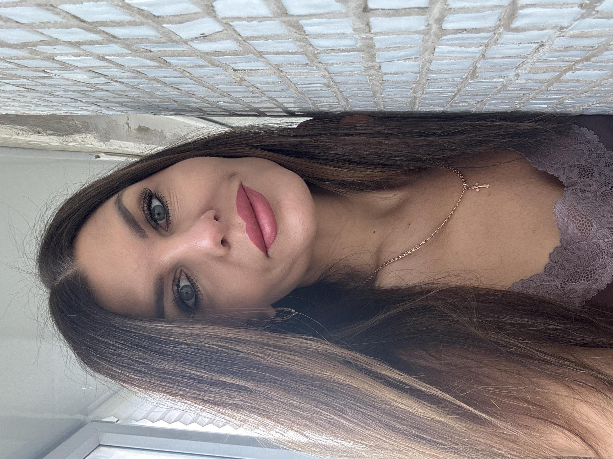 This media file features a portrait of a young woman with long, straight, dark brown hair that has lighter highlights. She gazes directly at the camera with green or hazel eyes and a neutral expression. Her lips are full and adorned with pink lipstick, and her eyebrows are well-defined. She is wearing a delicate gold-colored chain around her neck, which includes a small cross pendant. Part of a light purple, lace-trimmed top or camisole is visible on her right shoulder.
The background consists of a textured surface made of numerous small, light-colored square tiles, suggesting an interior wall or ceiling. Below this tiled surface and to the left of the woman, there is a hint of a lighter, possibly white, architectural element, which could be a window frame or sill, further indicating an indoor setting. The lighting appears to be natural, suggesting it is daytime. There is no visible text within the image. This indoor scene is located in Chaikovsky, Russia.
