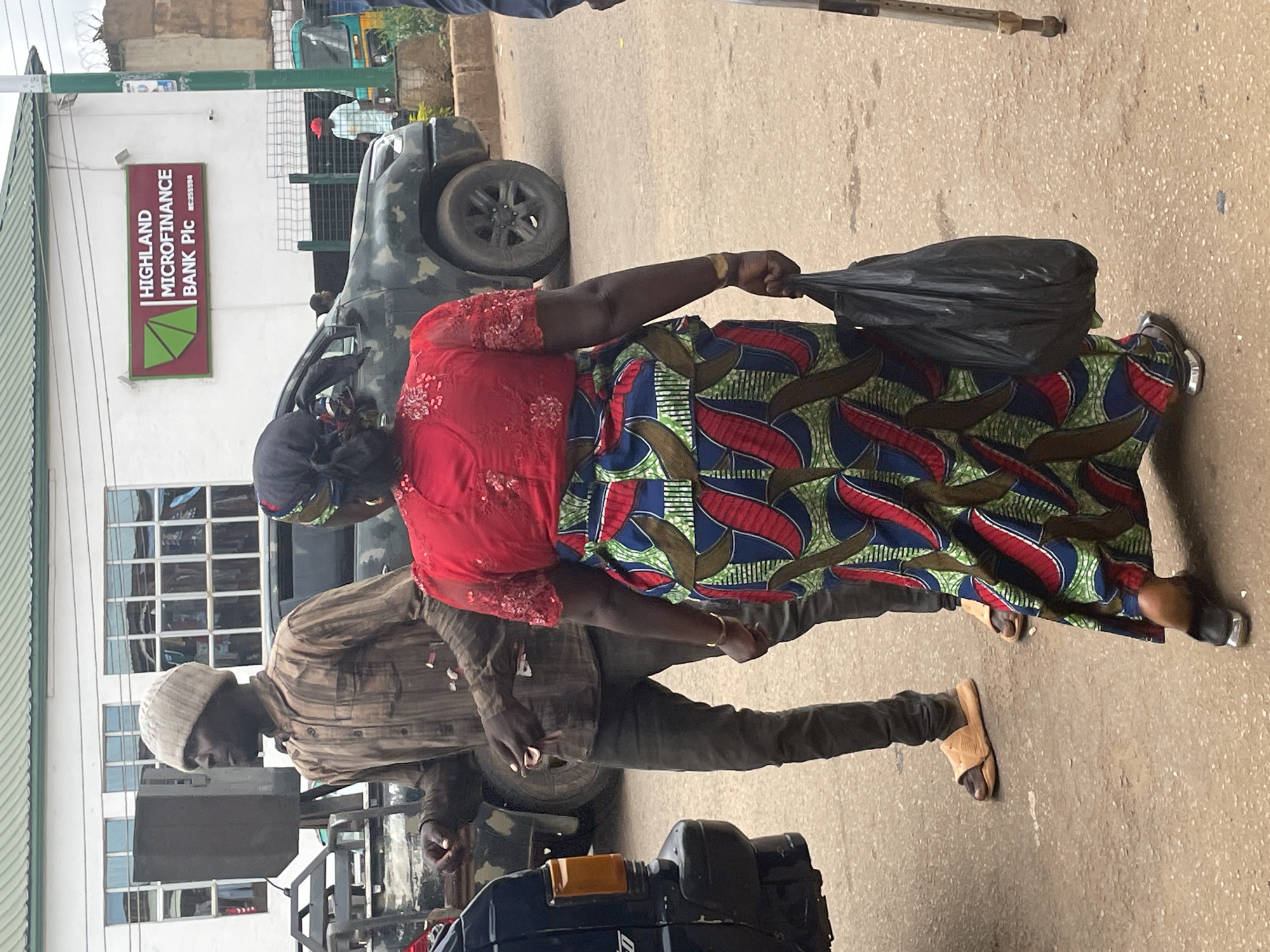 In Bukuru, Nigeria, a man wearing a brown striped long-sleeved shirt, dark trousers, and a light brown beanie hat stands near a black vehicle. He appears to be interacting with another person, though only their legs and feet are visible. To his right, a person wearing a vibrant red top with intricate beadwork and a multicolored patterned skirt is walking away, carrying a black plastic bag. They are both on a paved road, with a building in the background featuring a sign for "HIGHLAND MICROFINANCE BANK Plc." The environment suggests an urban setting, possibly a street in Bukuru.