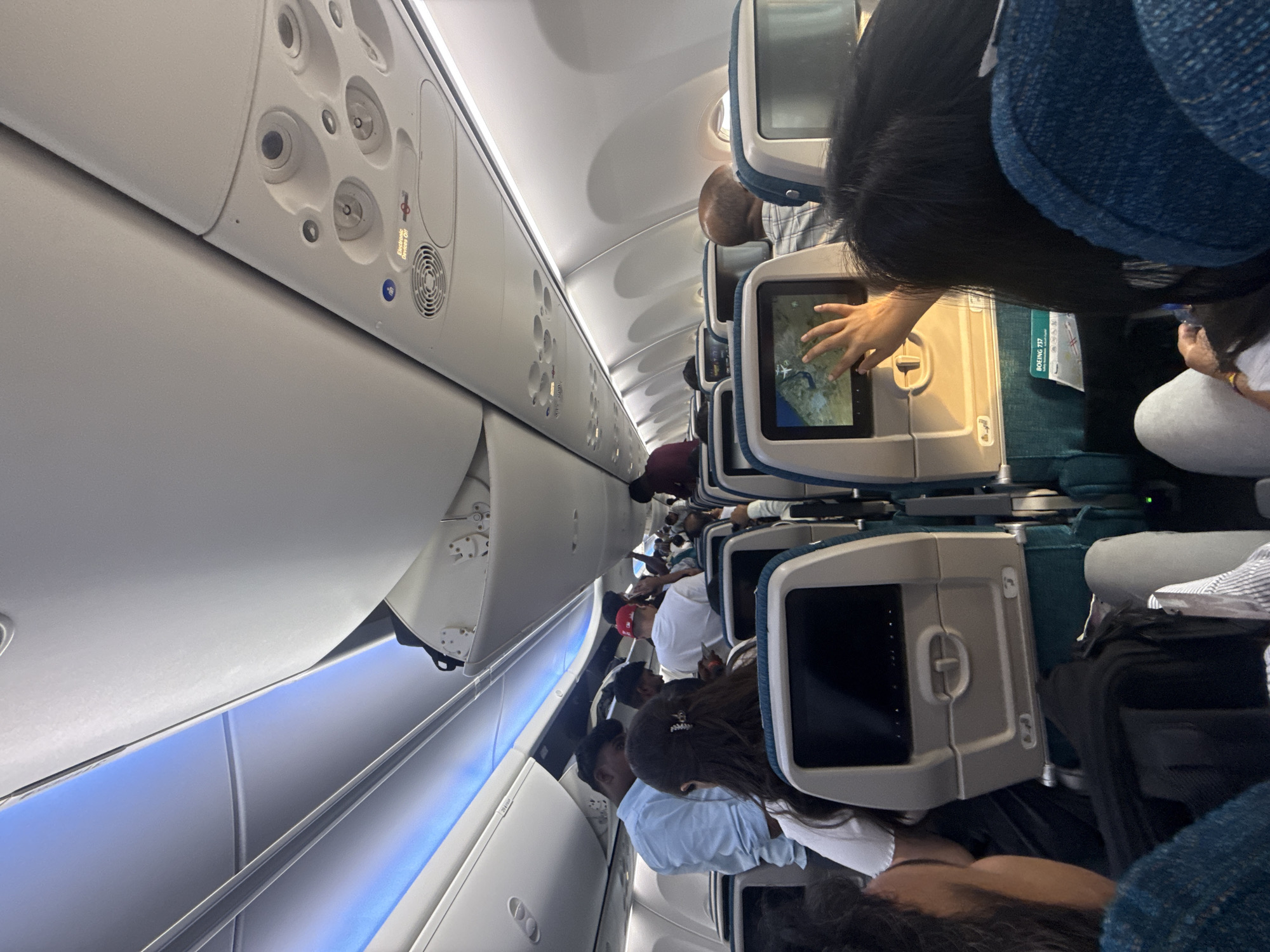 The image depicts the interior of an airplane cabin, with passengers seated and some standing in the aisle. The overhead compartments are visible, some of which are open. The seats have individual in-flight entertainment screens, one of which is being touched by a passenger's hand. A safety card for a Boeing 737 is visible on the seat back. The lighting in the cabin is a mix of overhead lights and blue accent lighting. The scene appears to be in motion, as suggested by the slight blur in some areas. The context provided suggests this image was taken in Dubai, United Arab Emirates, however there are no visual cues in the image that confirm this location.
