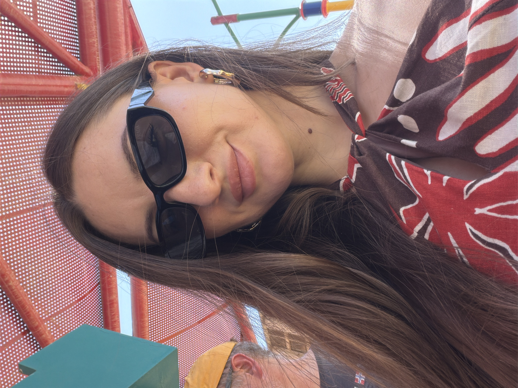 The image captures a close-up, waist-up selfie of a young woman outdoors, likely in a playground setting within Ash Sheikh Zayed, Egypt, indicated by the vibrant red, orange, and blue playground structures in the background. She is wearing black rectangular sunglasses and a brown and red patterned halter top that is tied at the front. Her long, brown hair is framing her face and falling over her shoulders. A man's head, wearing a yellow cap, is partially visible on the left side of the frame. The overall atmosphere is bright and sunny, suggesting a daytime activity.