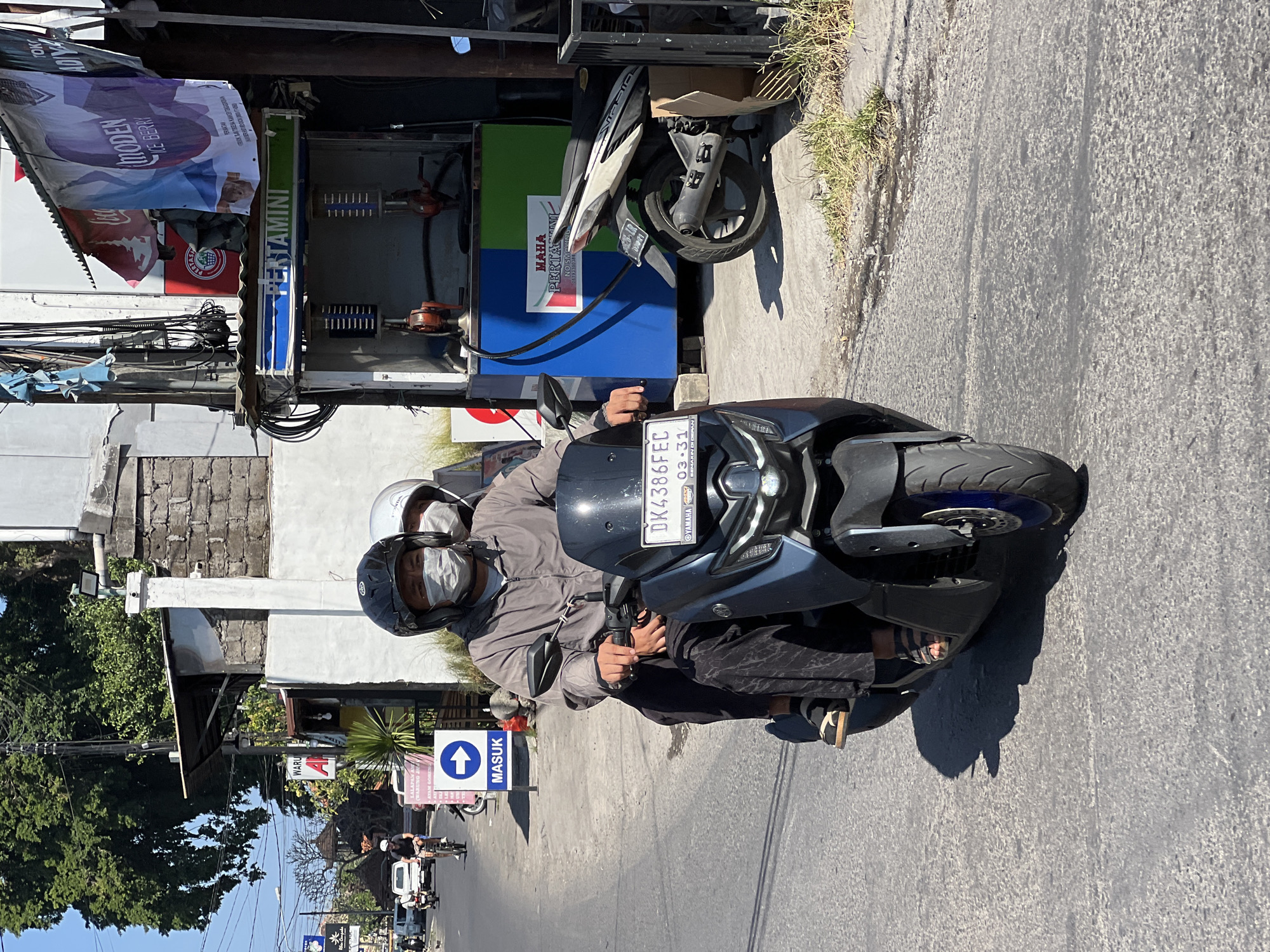 This is a medium-angle, eye-level, full-shot photograph of two people on a scooter on a sunny day in Denpasar, Indonesia. The scooter is dark gray and the rider in front is wearing a gray jacket and a black helmet with a white face mask. They are driving on a paved road. Behind the driver, a person wearing a white helmet and a gray face mask is visible. The background features a road with other vehicles, buildings, trees, and a "PERTAMINI" gas station. There's a blue and white sign with an arrow pointing to the right that says "MASUK." The license plate on the scooter reads "DK 4386 FEC."