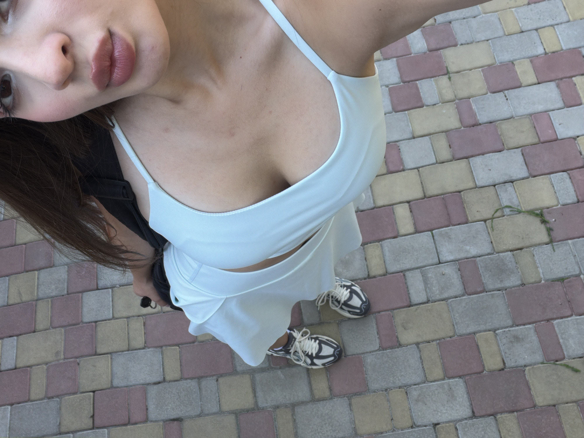A low-angle, eye-level, full shot shows a person from the chest down, wearing a pale blue slip dress and black and white sneakers. The person is standing on a paved walkway made of interlocking bricks in various shades of pink, yellow, and gray. The person's torso is visible, with the slip dress revealing cleavage. The person's legs are bent slightly as they stand. The background is a continuation of the brick walkway. The image is taken outdoors during the daytime, as indicated by the natural light.