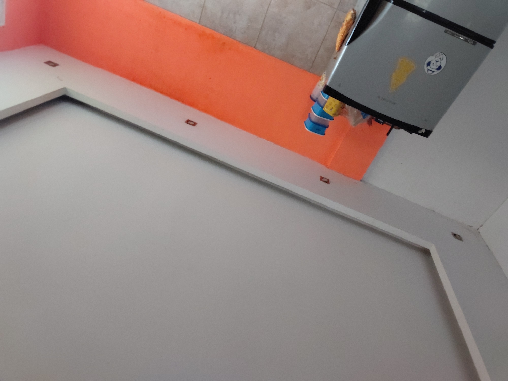 This image depicts an interior space, likely a kitchen or pantry area, characterized by a vibrant orange wall. The perspective is tilted, but when oriented correctly, a grey refrigerator is visible on the right, positioned against the orange wall. On top of the refrigerator, there's a baguette or loaf of bread and several stacked plastic containers, predominantly blue and yellow.
The refrigerator door features two stickers: a yellow, triangular shape, possibly a pizza slice or corn chip, and a circular white sticker with a blue border depicting a smiling cartoon character, possibly a chef or mascot. Faint text, which appears to be a brand name like "Tronex," is visible below the yellow sticker.
A white L-shaped surface, resembling a countertop or a deep shelf, extends along the orange wall, with small, square, brownish inlays embedded periodically. The background at the top reveals a light-colored tiled surface, suggesting either a backsplash or a tiled wall. Another wall to the left is painted a lighter shade, possibly white or light grey. The scene is devoid of people and shows no specific activity. The lighting suggests it is daytime. The provided context indicates the location is Santa Ana, Venezuela.
