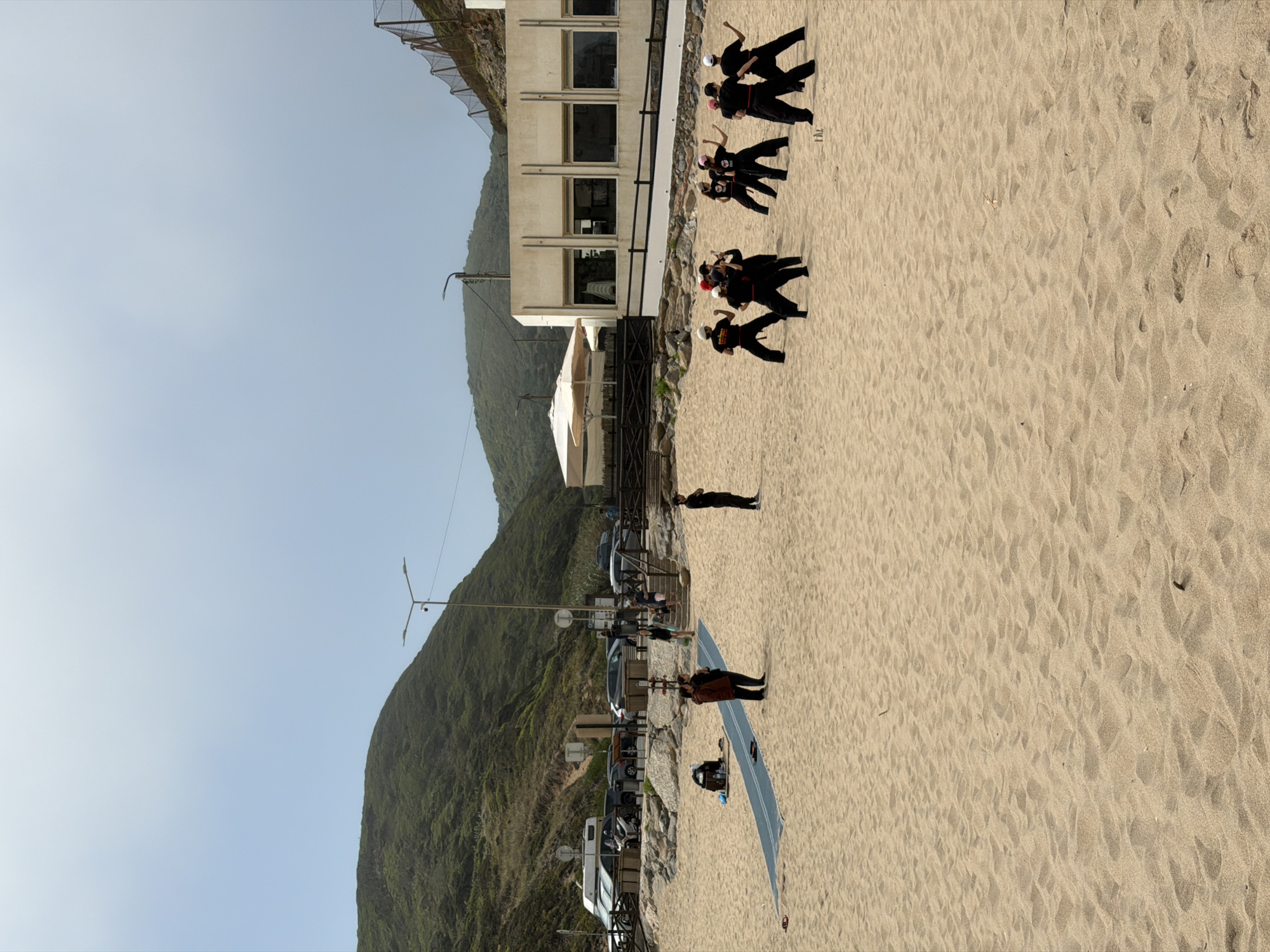 The image shows Praia das Maçãs, a beach located in Colares, Portugal. The scene captures a group of people practicing martial arts on the sandy beach. The group consists of several individuals, mostly young women, dressed in black martial arts uniforms and white or pink headbands, performing various stances and movements. In the background, there is a light-colored building with large windows, a white umbrella, and some parked vehicles, including a camper van. Further back, a lush green hillside forms the backdrop, with a road and a fence visible along its slope. The sky is overcast with a light haze. The sand on the beach has a textured surface with footprints and small debris. A blue mat is laid out on the sand in the foreground, with two people standing on it. Another individual is standing to the right of the mat, facing away from the camera.