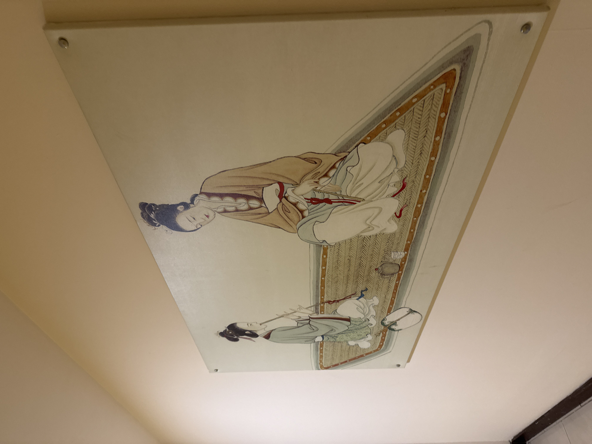 The image shows a framed artwork on a light beige wall. The artwork depicts two figures in traditional East Asian attire. The figure on the left, a woman with her hair in a bun, is playing a flute. She is wearing a grey robe with red trim and green patterned pants. She is seated on a woven mat. To her right, another woman, also with her hair styled up, is seated and appears to be holding a fan. She wears a tan robe over a white and light blue garment. They are positioned on a triangular mat with a decorative border. In the foreground, near the woman playing the flute, are two small ceramic objects that appear to be a teapot and a cup. The artwork is secured to the wall with visible silver pins at the corners. The wall itself is a muted yellow-beige. The lighting is soft, casting a slight shadow. There is a hint of a darker colored wall or trim along the bottom left edge of the image, suggesting a corner or doorway. The location in Aksu, Turkey is not discernible from the image itself.