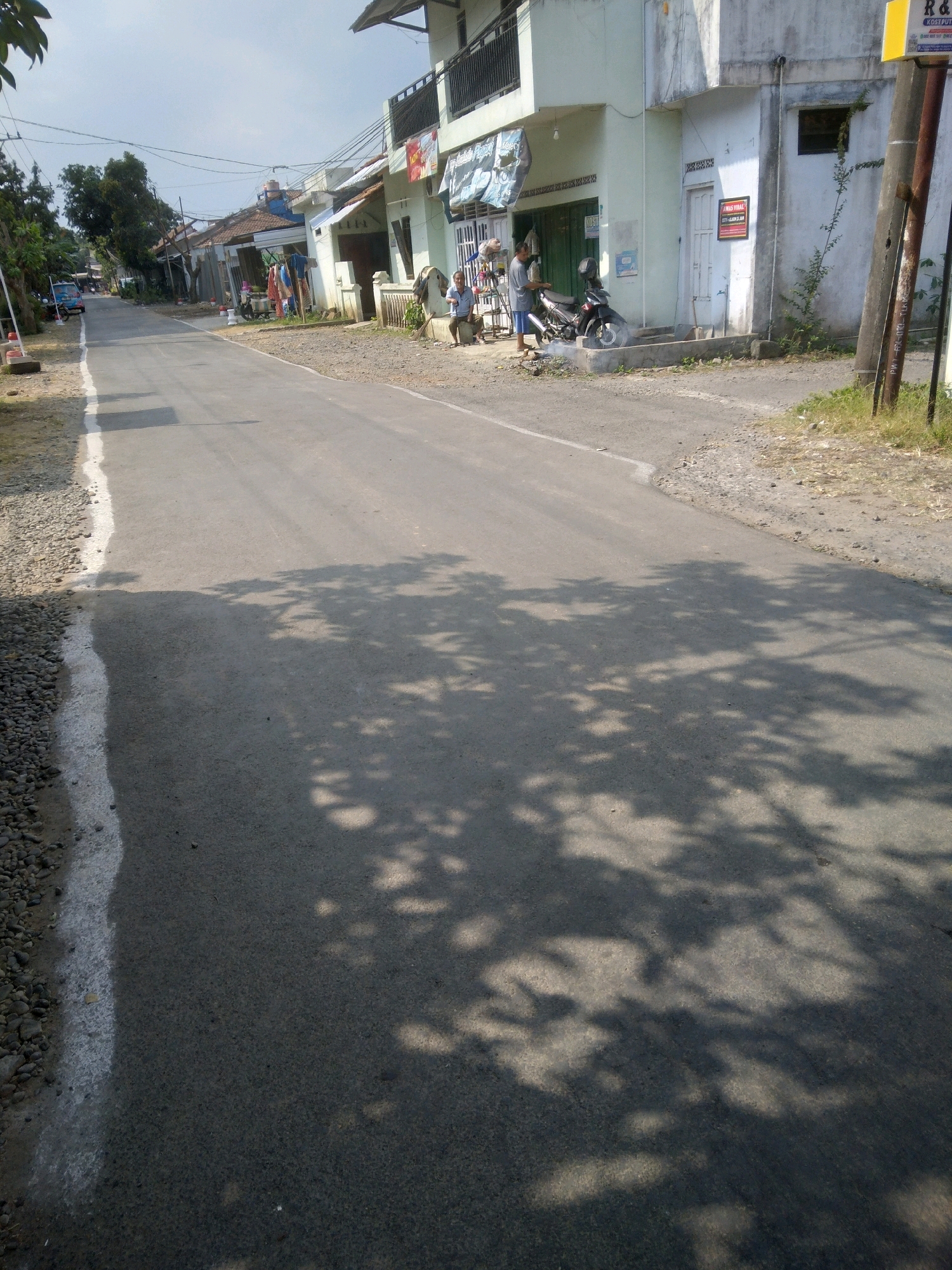 The image depicts a street scene in Desa Pamijen, Kecamatan Sokaraja, Indonesia, under a clear sky. The road, paved with asphalt and bordered by gravel and a white line on the left, stretches into the distance. On the right side of the road, several buildings are visible, characterized by their simple architecture and off-white facades. One building has a balcony with a dark railing. In front of one building, a motorcycle is parked on the pavement. A man in a grey shirt and blue shorts is standing near the motorcycle, appearing to interact with it. Another person is seated on a low object to the left of the man and the motorcycle. Further down the street on the left, laundry is hanging on a clothesline between two poles. Trees and power lines are present along the sides of the road. Shadows of tree leaves are cast across the asphalt in the foreground and midground, indicating the time of day. A sign with red and white lettering is affixed to one of the buildings. A yellow sign is also visible on a pole on the far right. The environment appears to be a residential or semi-commercial area with modest buildings and a rural or semi-urban feel.
