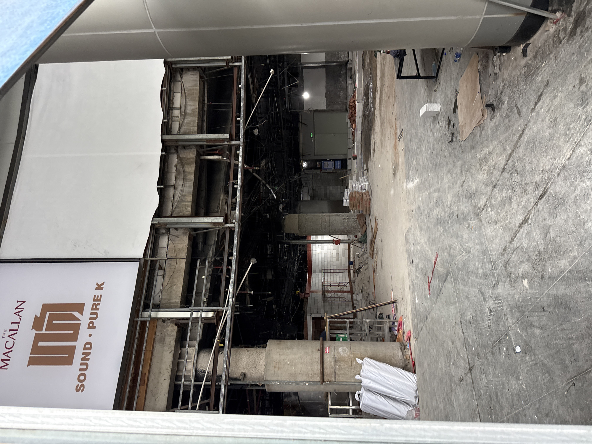 The image captures an interior view of a commercial space undergoing renovation or demolition in Tianhe District, China. The foreground is dominated by a concrete floor with visible grout lines and scattered debris, including a small white cylindrical object and a piece of cardboard. To the left, two large white bags are leaning against a substantial concrete pillar. Behind this, a metal scaffolding structure is partially visible.
The midground reveals the skeletal structure of the building, with exposed concrete beams, metal framing, and dangling wires and pipes. A large, imposing concrete pillar stands prominently on the left. Beyond this, the space opens up to reveal more construction materials, including stacks of bagged cement and piles of red bricks. In the distance, a set of double doors with an "EXIT" sign is visible, and a person wearing a yellow hard hat can be seen near the back wall, which is constructed of white cinder blocks.
The upper portion of the image features a large white sign with branding that includes "THE MACALLAN" and a logo with "SOUND • PURE K" below it. The surrounding architecture suggests a large, potentially public or commercial venue, with evidence of extensive construction work. The overall impression is one of an active construction site, with the remnants of past structures being replaced or renovated.