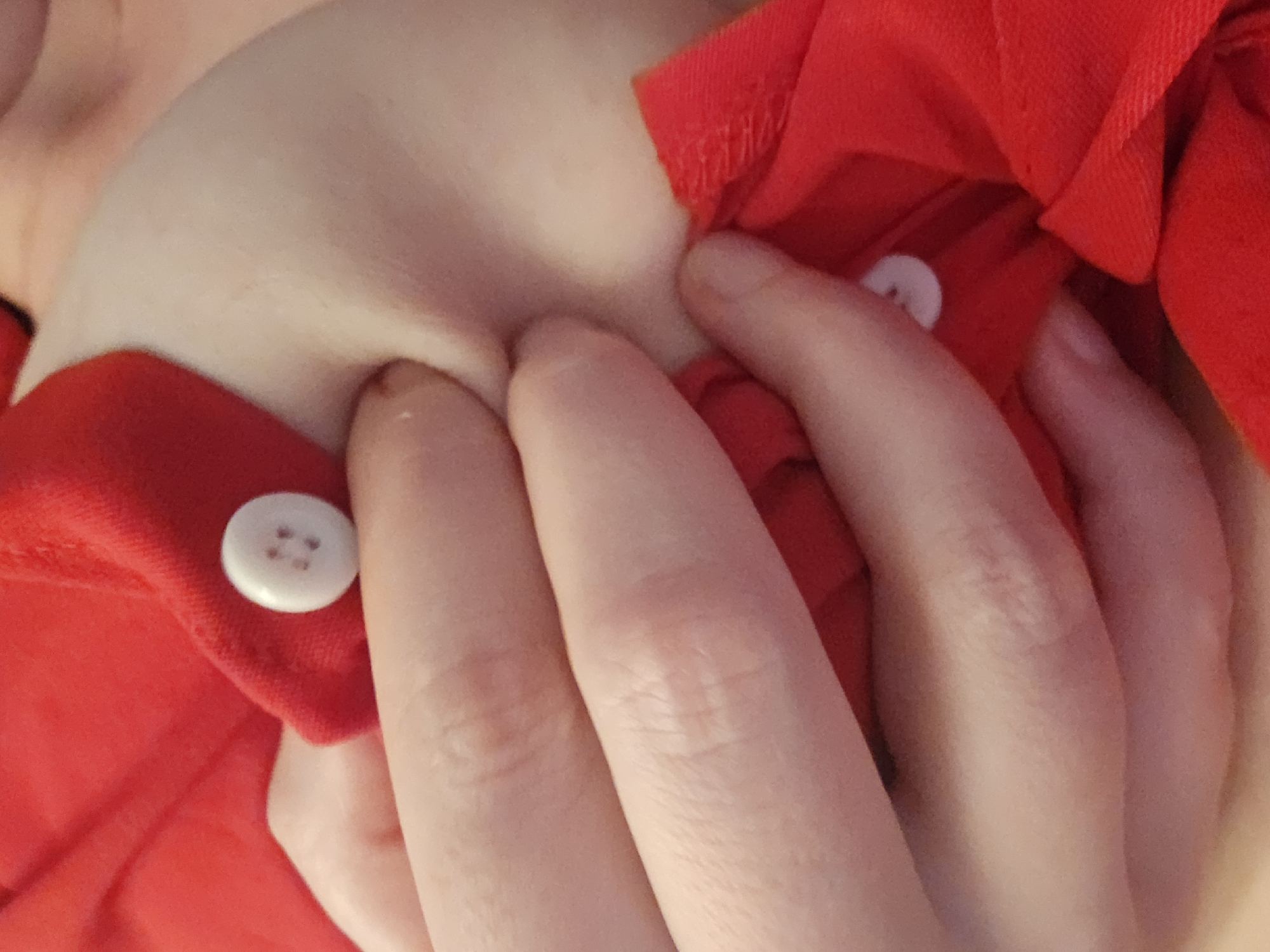 The media file shows a close-up, abstract view of a person's chest area, covered by a red garment. Two white buttons are visible on the fabric. The person's fingers are interlaced and pressing into their chest, obscuring most of the view. The lighting suggests an indoor setting, possibly with soft, ambient light. The overall impression is one of intimacy or personal reflection, with the focus on the texture of the fabric, the skin, and the human form. There is no discernible text in the image. The activity appears to be a self-touching or resting gesture, with the fingers creating shadows and folds in the red material. The image lacks any external cues about the time of day, weather, or specific location.