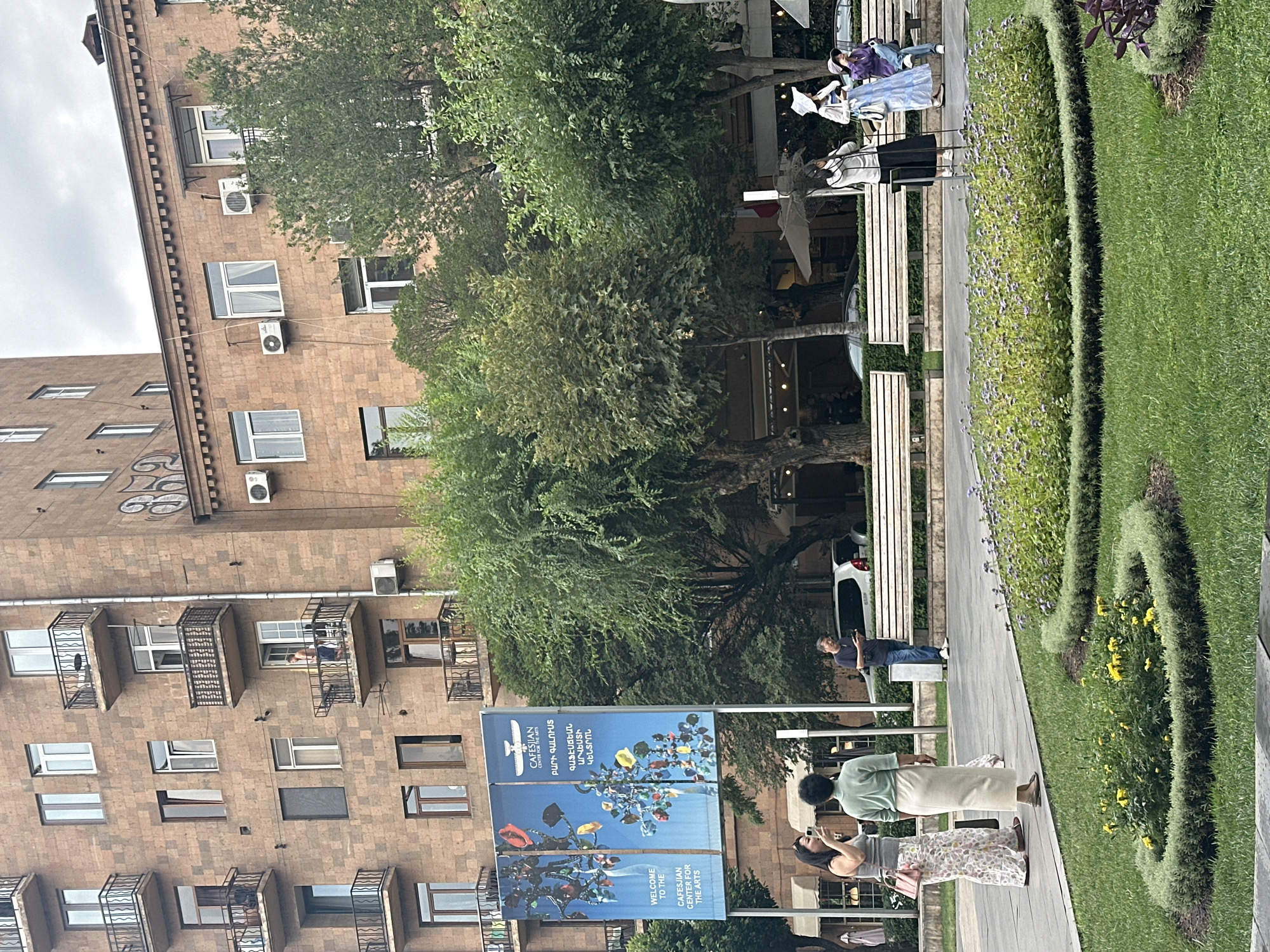 The image depicts a street scene in Yerevan, Armenia, likely near the Cafesjian Center for the Arts, as indicated by a banner advertising it. In the foreground, a manicured garden with blooming yellow and purple flowers is visible, set against a backdrop of green grass. A paved walkway runs alongside the garden, where several individuals are present. On the left, two women stand on the walkway, one wearing a patterned skirt and a grey top, the other in a light green skirt and top. To the right, a man in a dark t-shirt and jeans stands looking at his phone. Further back, a white car is parked, and a series of park benches line the area. In the mid-ground, a multi-story brick building with numerous balconies and windows dominates the scene. Trees with lush green foliage are interspersed between the building and the street. The sky is overcast, suggesting a cloudy day.