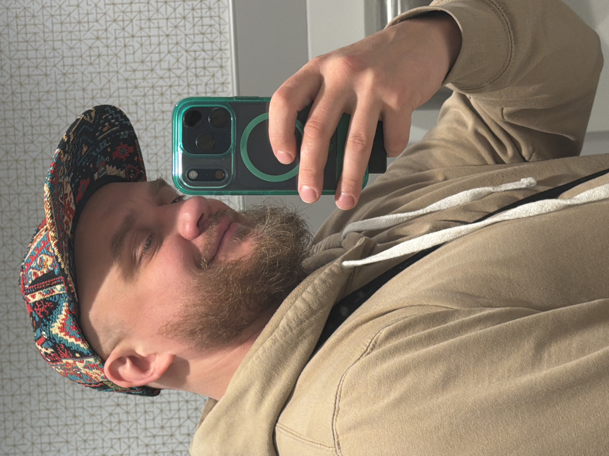 A man takes a selfie in front of a patterned wall. He wears a multi-colored hat and a tan hoodie with white drawstrings. He holds a smartphone with a green case in his right hand, angled towards his face. The background features a light-colored wall with a geometric pattern. The photo is well-lit, highlighting details of the man's face and clothing. No indication that the photo was taken in Chicago is present.