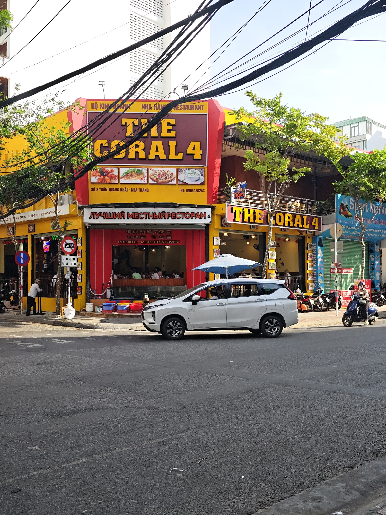 This is a street-level, eye-level, full shot of a building in Nha Trang, Vietnam, housing multiple businesses. The primary focus is on "THE CORAL 4" restaurant, a seafood establishment with a vibrant yellow facade and a maroon awning displaying the restaurant name in large, yellow lettering. Beneath the awning, the restaurant's interior is visible, featuring tables and patrons. Three framed images of seafood dishes are displayed on the yellow section of the building above the restaurant entrance. To the left of "THE CORAL 4," another establishment with a yellow facade is visible, exhibiting an open doorway with a person standing inside. A blue and white circular sign with a diagonal red line is mounted on a pole near this entrance. To the right of "THE CORAL 4," a "Sport Market" store is situated, with a closed blue storefront. The street in front of the buildings is paved and shows signs of wear. Several motorcycles and scooters are parked along the sidewalk. A white car is driving in the foreground, obscuring part of the sidewalk and the building's lower section. Overhead, a dense network of black electrical wires crisscrosses the frame against a bright, clear sky. Trees with green foliage are interspersed along the building fronts.