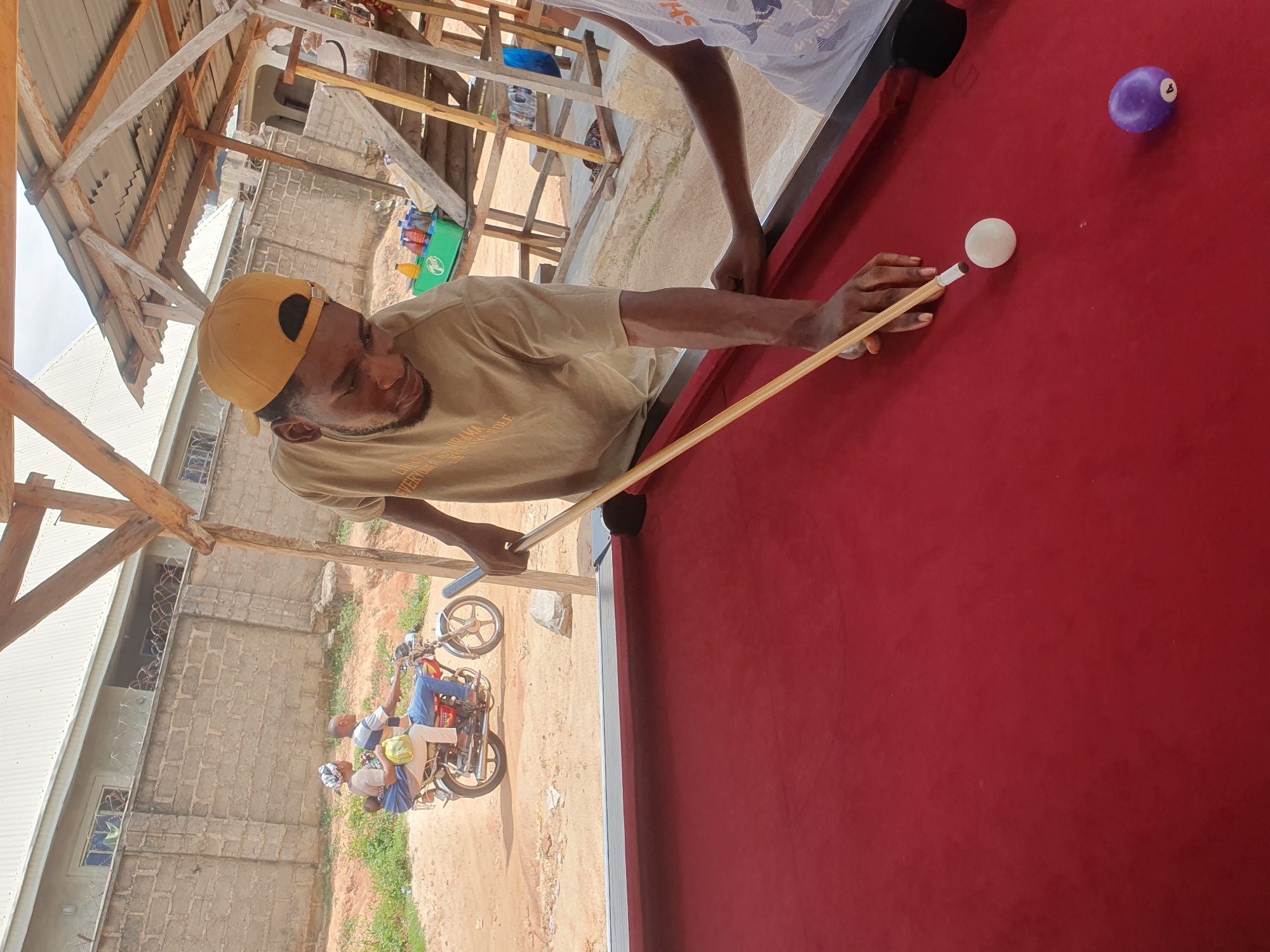 In Lokoja, Nigeria, a man wearing a yellow baseball cap and a light brown t-shirt is playing pool. He is positioned on the right side of a red pool table, holding a pool cue and preparing to strike a white ball. A purple billiard ball with the number 4 is visible on the table to the right of the white ball. In the background, a man and woman are riding a motorcycle, with the woman carrying a baby. The environment appears to be an outdoor recreational area with a simple wooden structure providing shade, and a brick wall and a building are visible further in the background. The ground is dusty and unpaved.