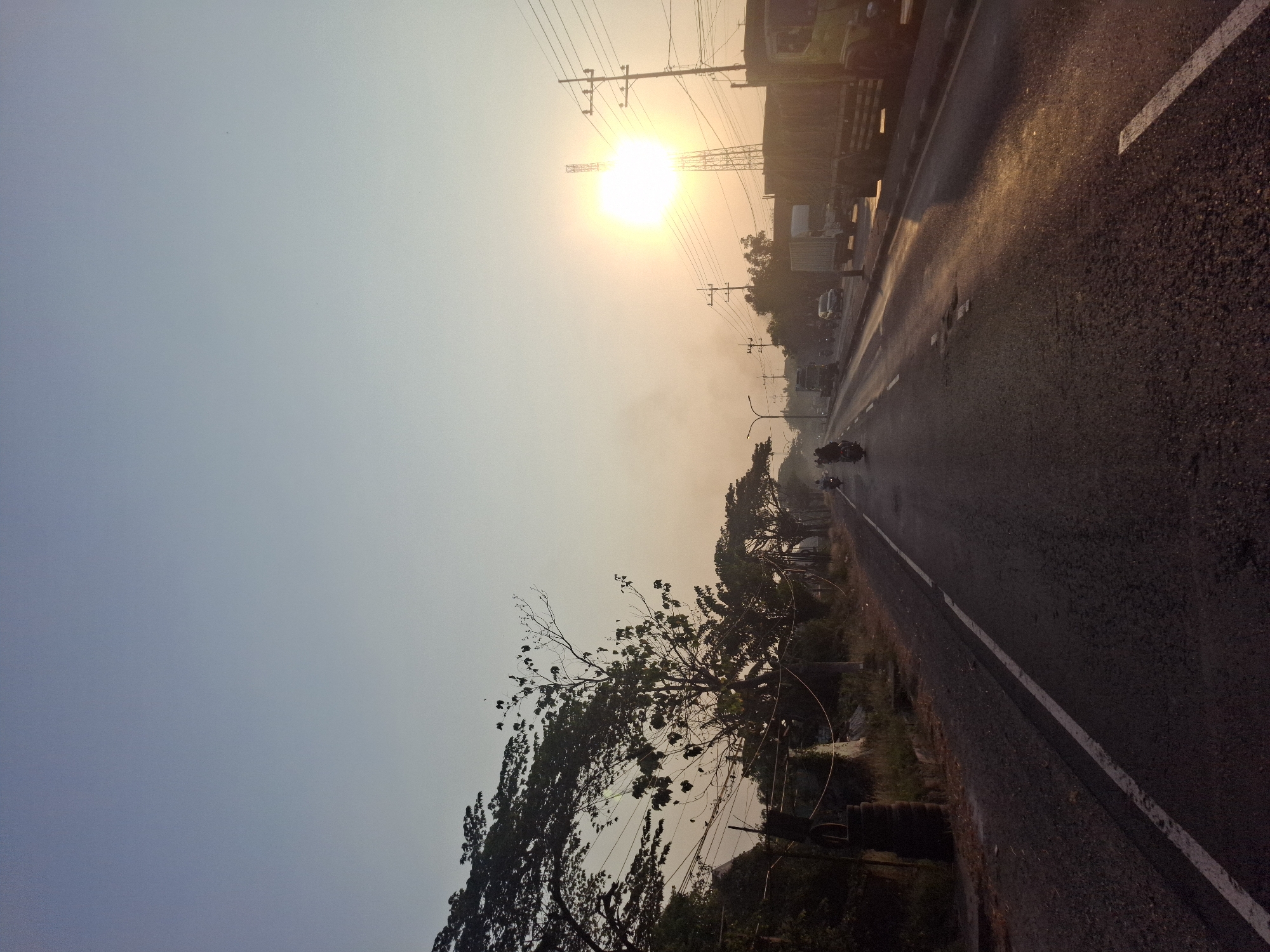 The image depicts a road in the city of Waruwetan, Indonesia, at sunrise. The sky is clear and pale blue, transitioning to a light orange hue near the horizon. The sun, a bright, almost blinding disc, is positioned in the upper right quadrant of the image, partially obscured by a tall transmission tower. The sunlight reflects off the wet asphalt road, creating streaks of golden light.
To the left of the road, a line of trees with dark foliage runs along the embankment, with some branches extending over the road. Telephone poles and power lines are visible throughout the scene. On the right side of the road, several trucks are parked or moving slowly, their cargo areas mostly obscured.
Several motorcycles and a few cars are traveling on the road. The motorcycles appear to be carrying one or two riders. The overall atmosphere is calm and serene, characteristic of early morning traffic in a developing region. The road surface is asphalt with visible white lane markings. The embankment on the left has some debris and vegetation. The image captures a typical scene of daily life and transportation in a city during the early hours of the day.