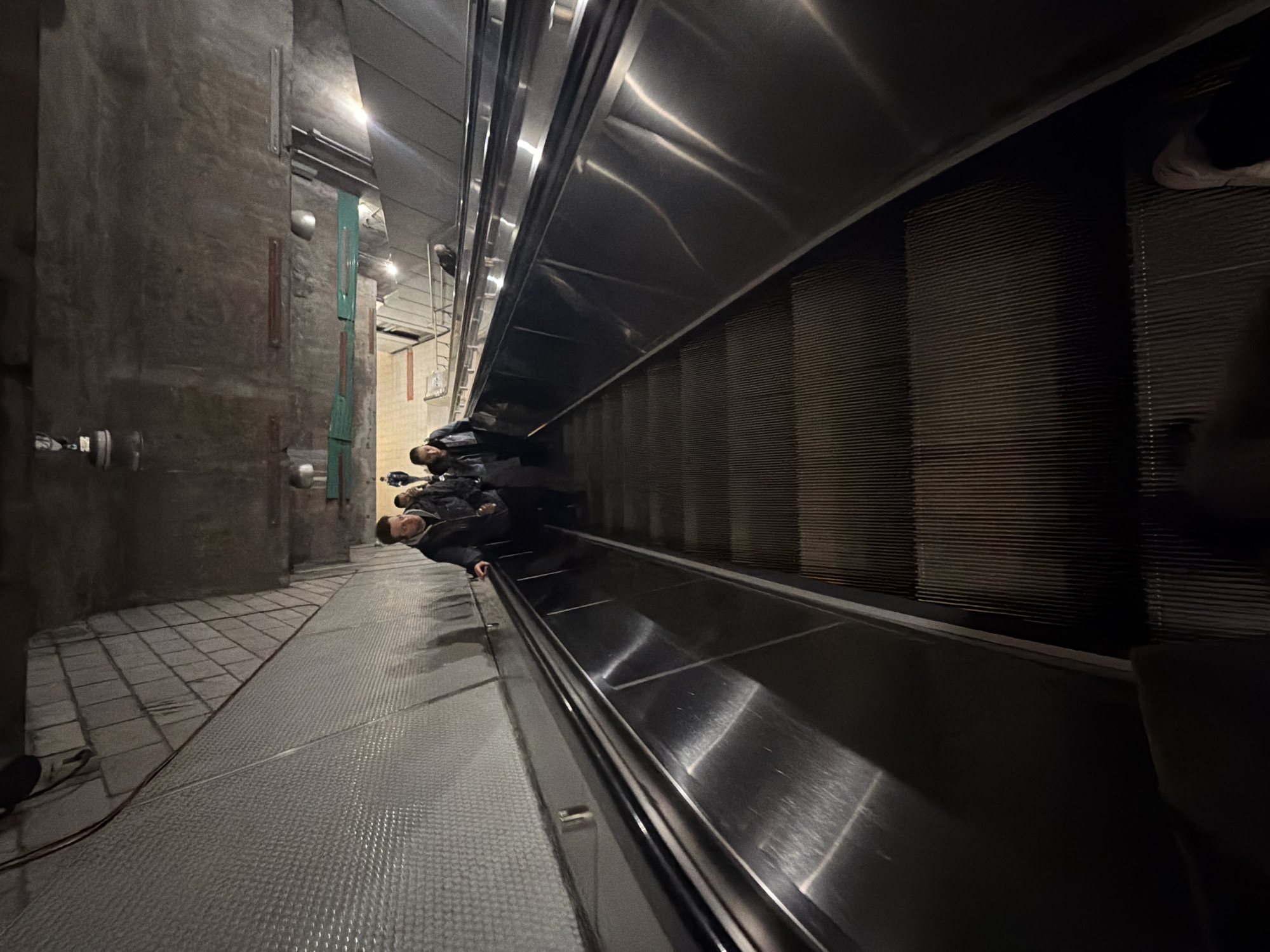 A low-angle perspective depicts an escalator in a transit station. The escalator is descending, with its metallic steps and side panels prominently displayed. Several individuals are using the escalator. The station has a utilitarian design with gray concrete walls and a metal floor. The photograph was taken in Montreal, Canada.