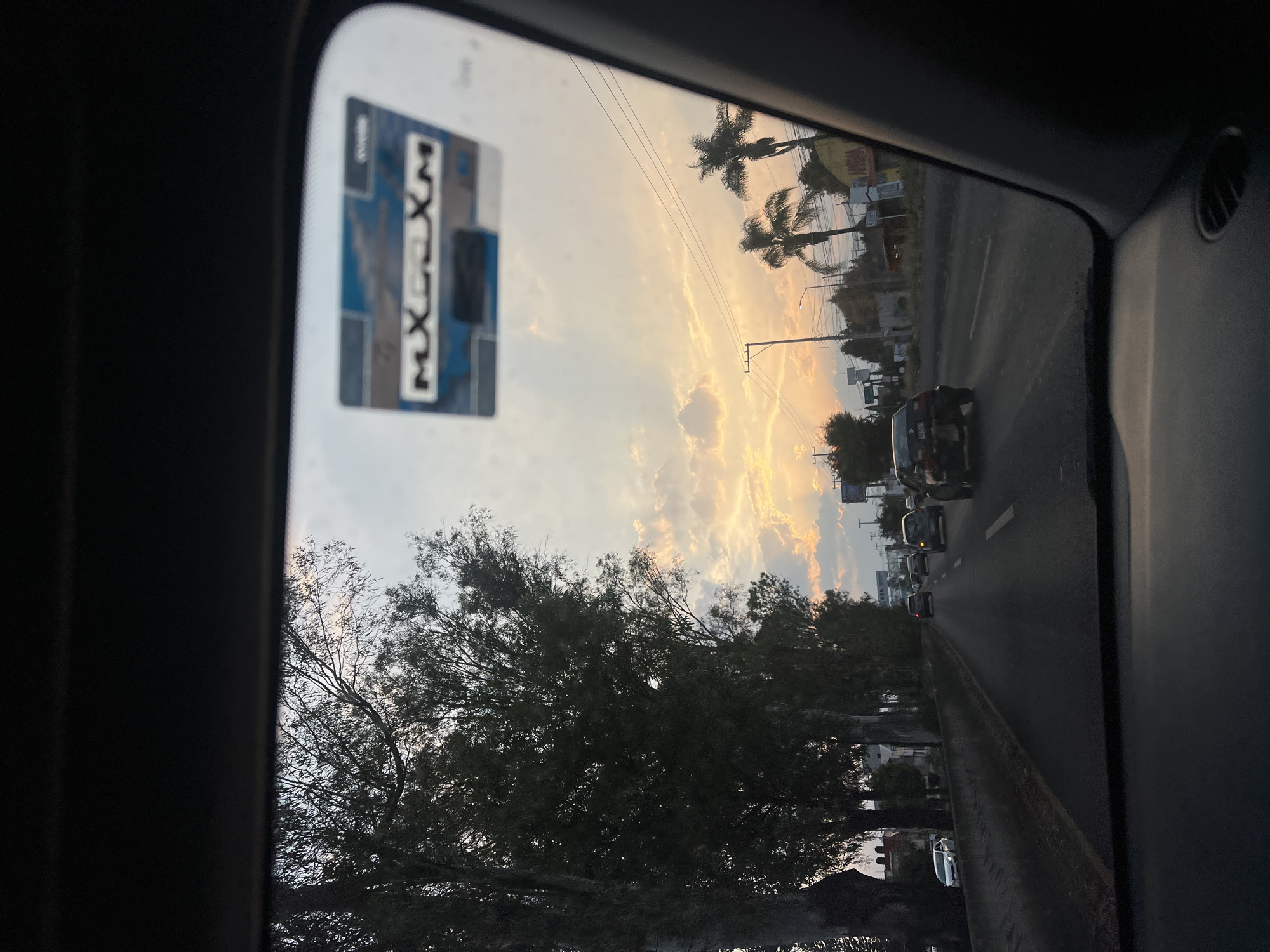 The photograph is taken from inside a car in Puebla, Mexico, looking out the windshield. A highway is visible with several vehicles. There are trees and palm trees visible along the roadside. The sky is a mix of clouds and sunlight. There is a parking pass in the upper left corner of the windshield.