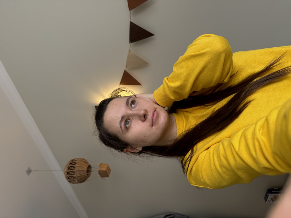 A young adult female, with long dark hair and a light complexion, is depicted from the chest up, wearing a bright yellow long-sleeved sweatshirt. Her left hand is raised, resting against the side of her head near her ear, and her gaze is directed upwards and to the right, exhibiting a neutral, contemplative expression. The indoor background features light-colored walls. A wicker hot air balloon decoration hangs from the ceiling in the upper left. Along the upper right wall, a string of triangular pennants in varying shades of brown and beige is visible. A warm light source illuminates the wall behind the subject's head. White crown molding is present at the junction of the wall and ceiling. A partial view of a dark rectangular object is visible in the lower left corner. The scene is located in a room in the city of Odintsovo, Russia.