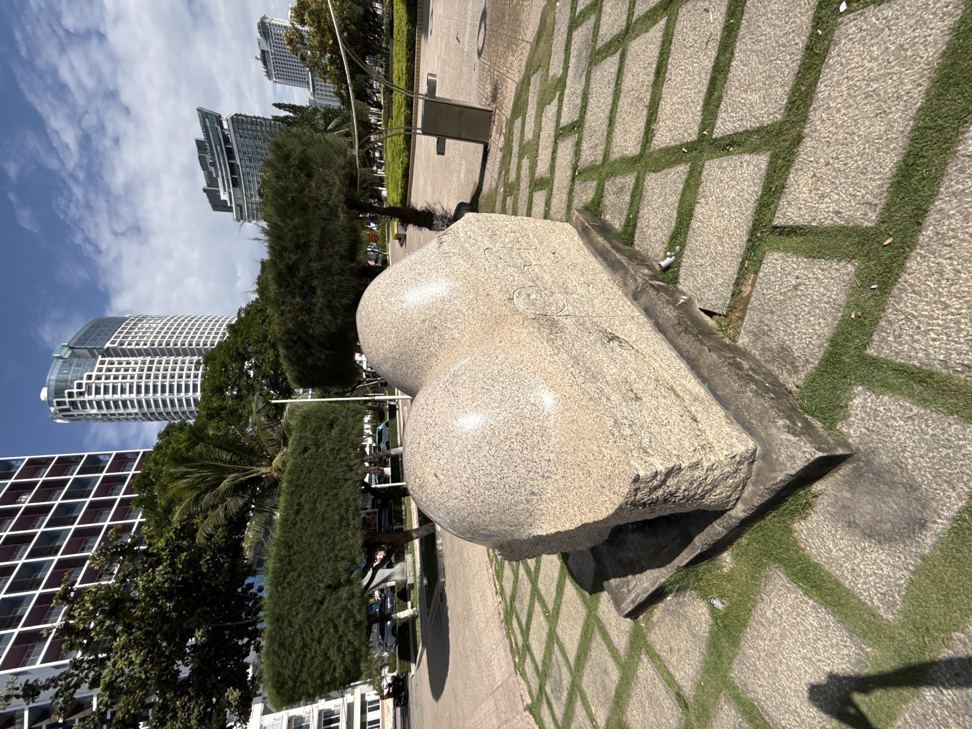 A low-angle, full shot captures a stone sculpture in the foreground, with modern high-rise buildings and a blue sky with scattered clouds in the background. The sculpture, made of speckled granite, has a rounded, bulbous shape with visible rough-hewn edges and some carved markings on its side. It is situated on a square concrete base set into a paved walkway with alternating square sections of grass. The walkway is patterned with diamond shapes formed by the intersecting lines of the paving stones and grass.
In the middle ground, manicured, rectangular bushes flank a paved area with trees and a street visible beyond. Cars and a motorcycle are parked or moving on the street. A lamp post stands to the right of the bushes. In the background, a cluster of contemporary high-rise buildings with distinctive architectural features dominates the skyline, suggesting an urban coastal environment. The architecture of the buildings, particularly the patterned facades and curved glass elements, points to the city of Nha Trang, Vietnam. The lighting indicates it is a sunny day.