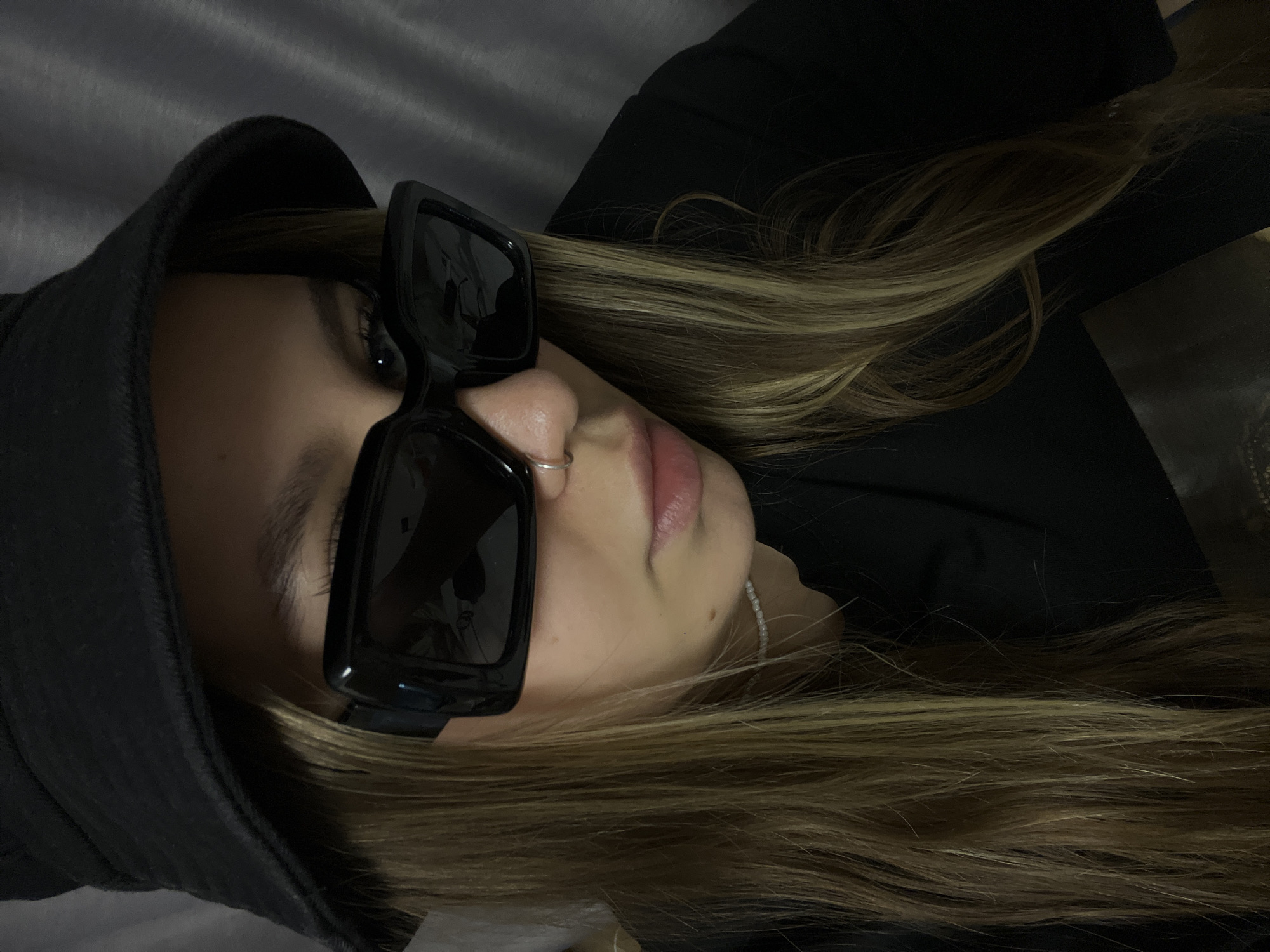 The image features a close-up portrait of a person, likely a woman, looking directly at the camera. She wears large, black, square-framed sunglasses that cover her eyes. A dark, possibly black, bucket hat or similar headwear covers the top of her head. Her long, light brown or blonde hair, appearing straight or slightly wavy, cascades over her shoulders and chest. A small silver hoop nose ring is visible on her left nostril, and a thin, white beaded necklace adorns her neck. Her lips are slightly parted, showing a natural lip color. She is wearing dark clothing, possibly a jacket or top, which is partially visible. The background is uniformly dark and appears to be a textured fabric, possibly a curtain or upholstered wall. The overall lighting is dim, creating a somewhat moody or stylish atmosphere. The person appears to be posing for the photograph. There is no visible text in the image. This scene takes place in Krasnodar, Russia.