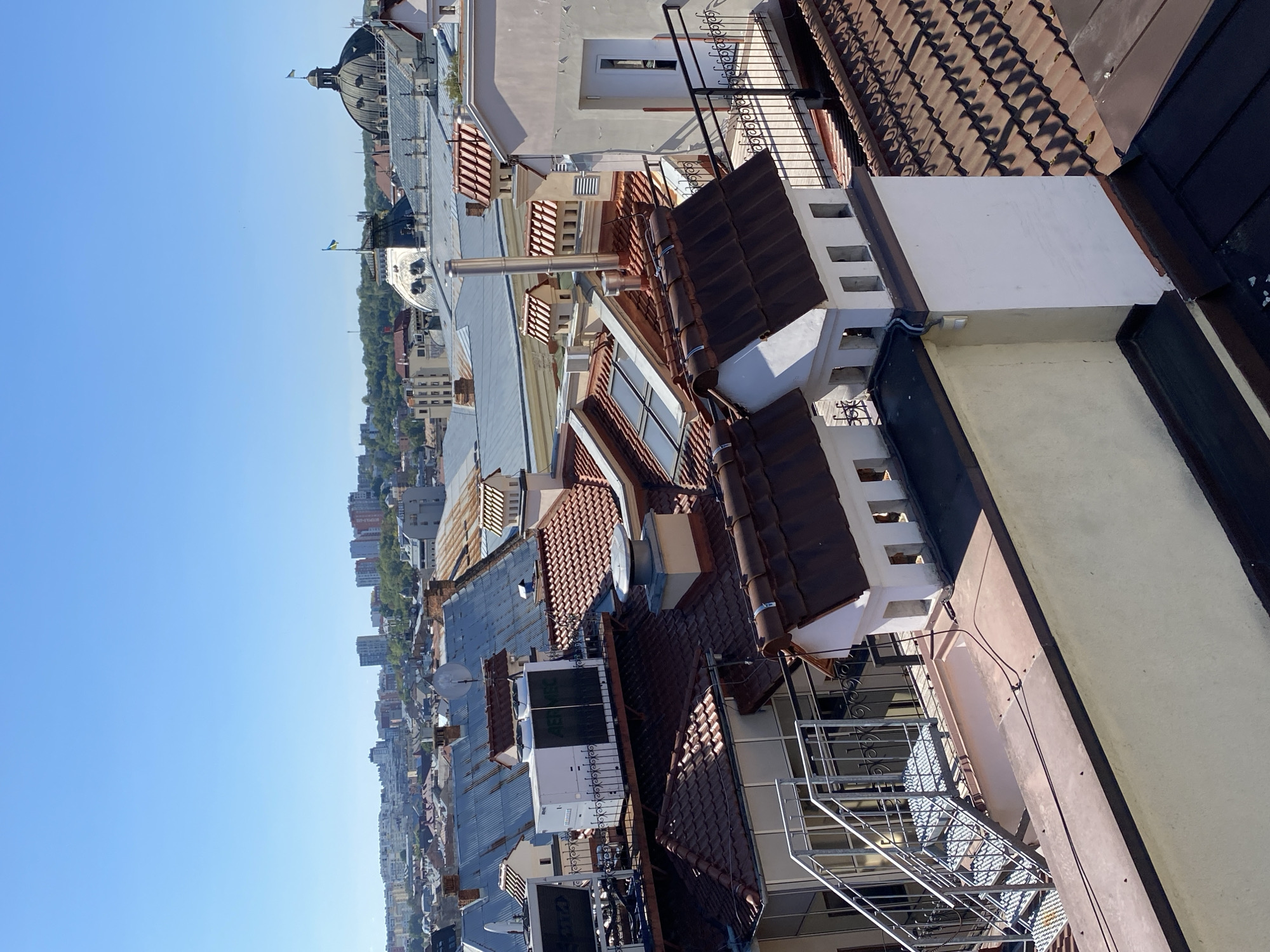 An aerial view captures a dense urban landscape of Lviv, Ukraine, dominated by a mosaic of rooftops under a clear blue sky. The foreground features a prominent cream-colored building with a white door and a black railing on a small balcony. Adjacent to it, a series of tan-colored, gable-roofed structures with decorative white chimneys and brown tiled roofs are visible, some with open rectangular window-like openings. To the left, a metallic outdoor staircase with diamond-patterned treads leads down to a flat roof.
Further into the mid-ground, a variety of rooftops are seen, including weathered metal roofs, red-tiled roofs, and some with modern air conditioning units. A large, white HVAC unit with "AER MEC" branding is a notable object on one of these elevated platforms, secured by a dark metal railing. Another prominent feature in the mid-ground is a large, dark dome topped with a Ukrainian flag. Two other Ukrainian flags are also visible on other buildings, one atop a white, stepped roof structure and another near the dome.
The background reveals a cityscape with numerous multi-story buildings of varying architectural styles and heights, interspersed with greenery. The overall scene suggests a sunny day in a historic European city, with a blend of older and more modern architecture. The vantage point is from a height, looking out over the urban fabric.