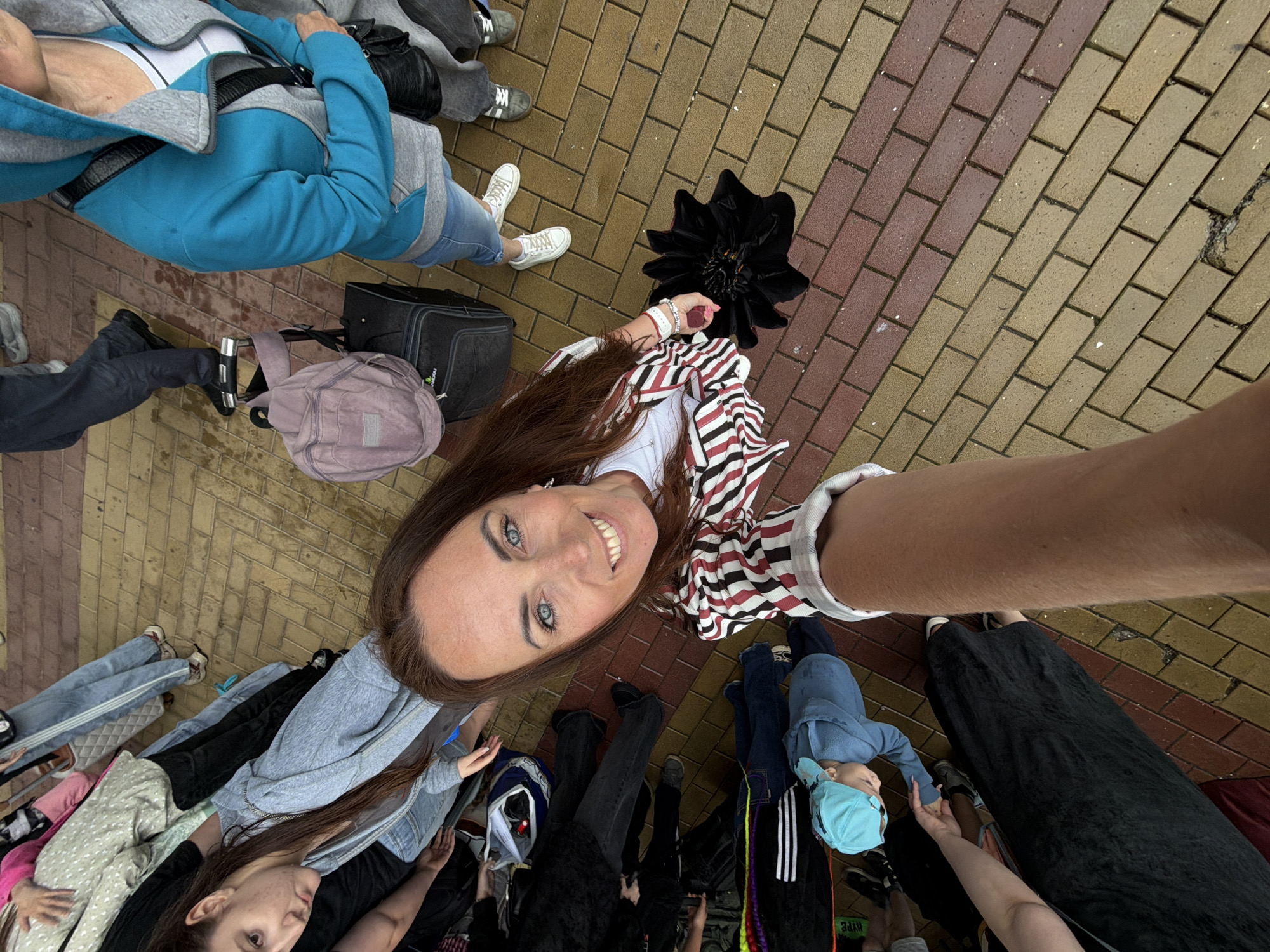 The image is a high-angle shot of a group of people on a brick-paved outdoor area, possibly a pedestrian walkway or plaza, in Birobidzhan, Russia. In the foreground, a woman with brown hair is taking a selfie, smiling at the camera. She is wearing a striped jacket over a white top. To her right, a black umbrella is lying open on the ground. Other people are visible in the background, some standing and others sitting or lying on the ground. One person, a child wearing a blue hat, is lying on the ground. Several pieces of luggage are also present, including a rolling suitcase and a backpack. The paving stones are a mixture of tan and reddish-brown bricks. The overall lighting suggests it is daytime, with some overcast conditions. The scene appears to be a public gathering or event.