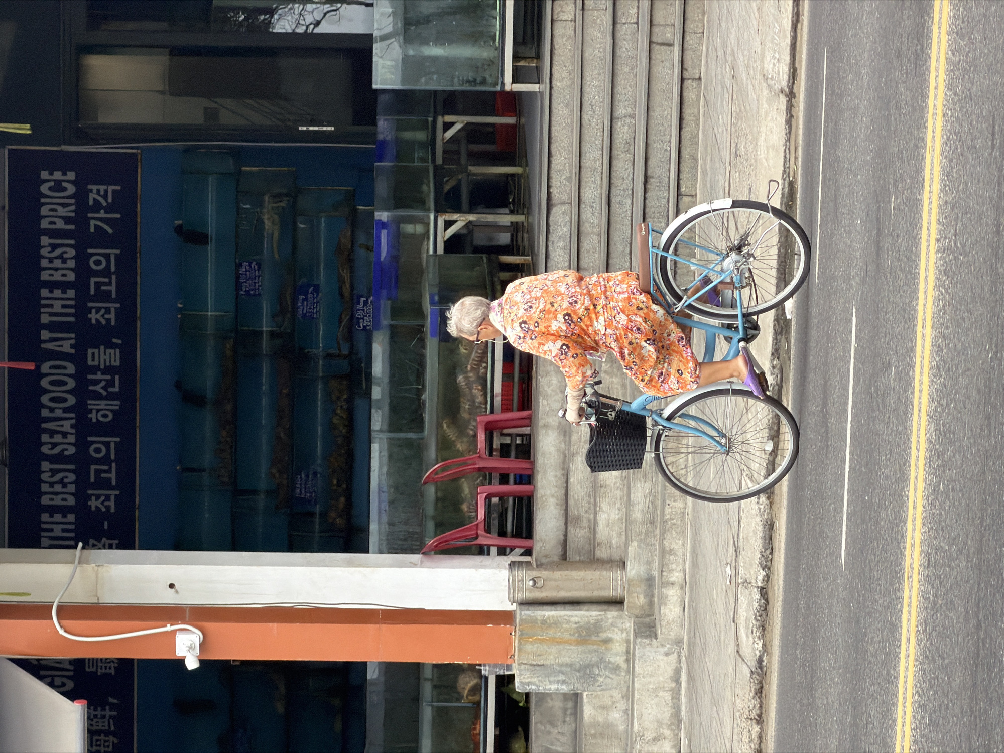 A senior woman with gray hair is riding a light blue bicycle with a black basket on a concrete sidewalk next to a street with yellow lines. The woman is wearing an orange floral dress and lilac-colored gloves. Behind her is a building with a sign in Korean that reads "THE BEST SEAFOOD AT THE BEST PRICE.” In the foreground are red plastic chairs and stacked glass tanks. Based on the Korean signage, this image is located in the city of Đà Nẵng, Vietnam.