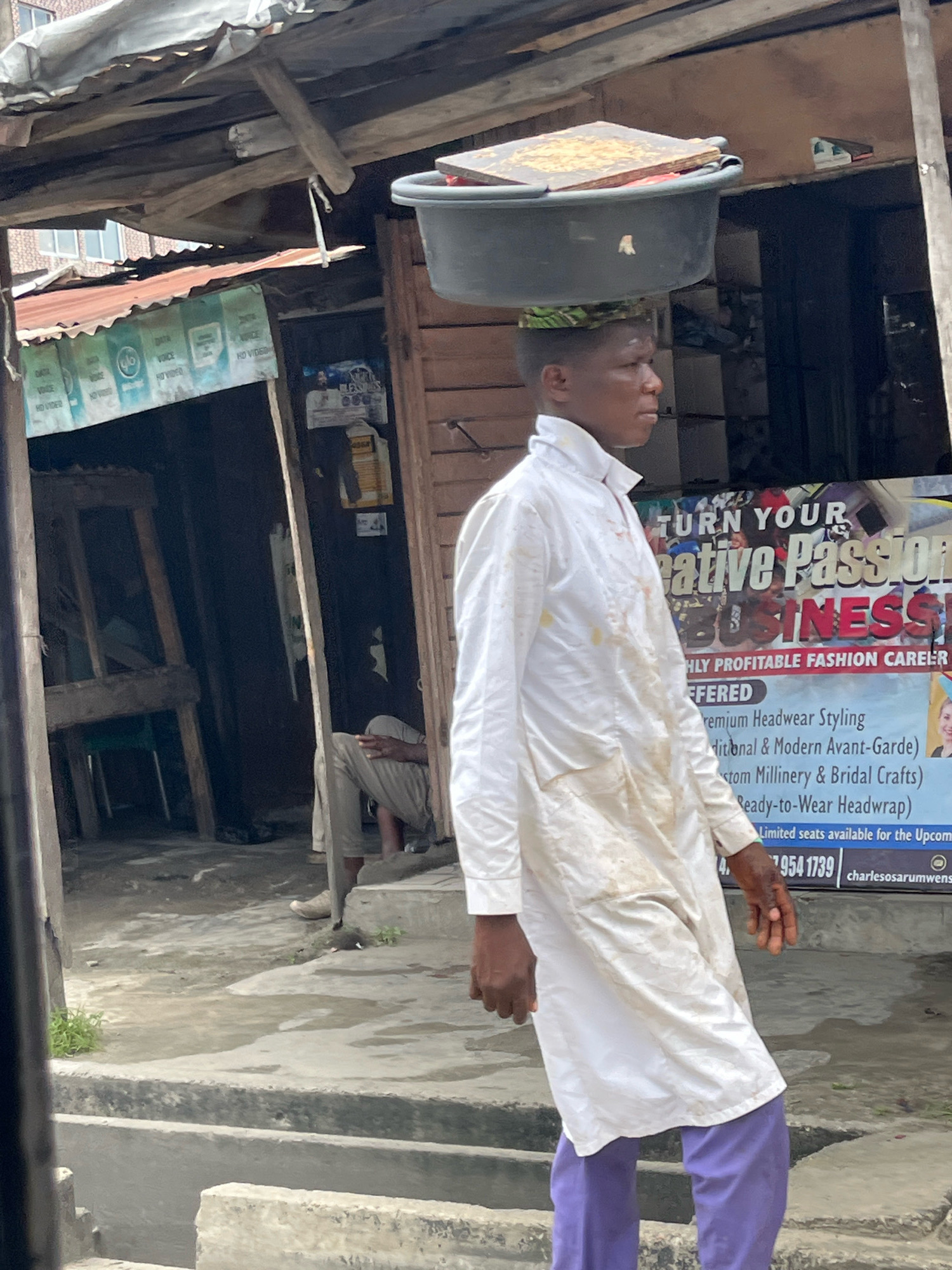 A young Black male in Ado, Nigeria, walks with a large grey basin filled with goods balanced on his head. He is wearing a white traditional Nigerian outfit and purple pants. The man is walking past a shop with a corrugated metal roof and wooden walls. Behind him, a sign advertises "TURN YOUR CREATIVE PASSION BUSINESS." A person is seated inside the shop, visible through an open doorway. The ground is paved with concrete and shows signs of wear and puddles of water. Green vegetation is growing in patches on the ground and along the edge of a drainage channel.