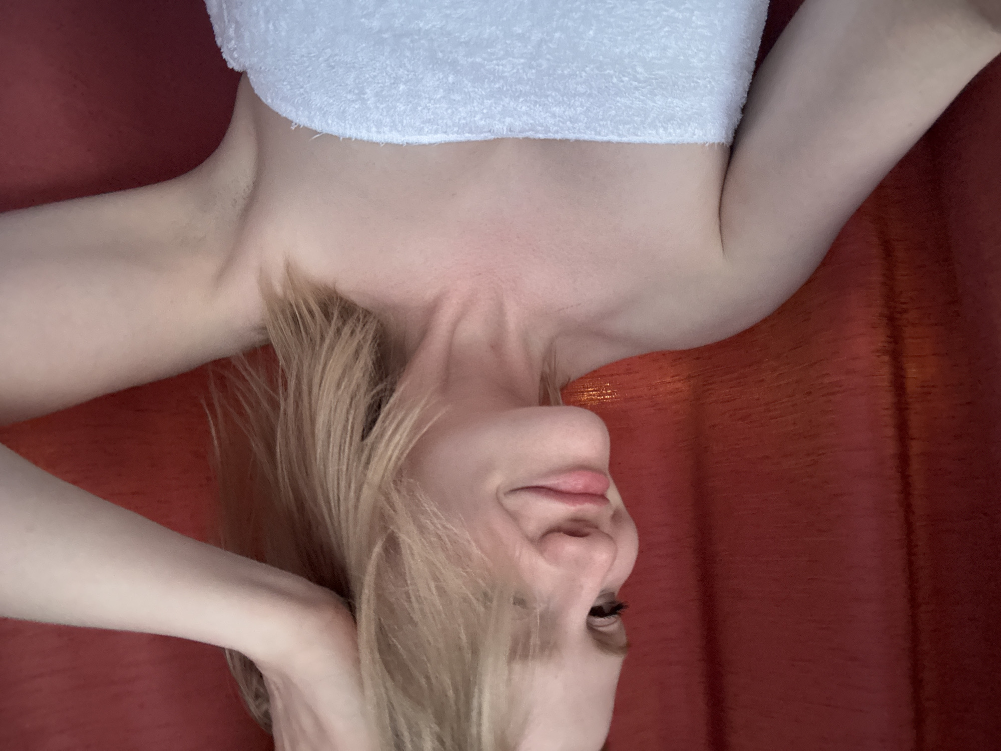 An individual with fair skin and blonde hair is positioned in front of a red curtain. The individual is wearing a white towel wrapped around their chest. Their left arm is raised, with their hand touching their hair. The individual is looking to the left and smiling slightly. The background is a red curtain, which appears to be textured. There is no information in the image that indicates the location in Saint Petersburg, Russia.