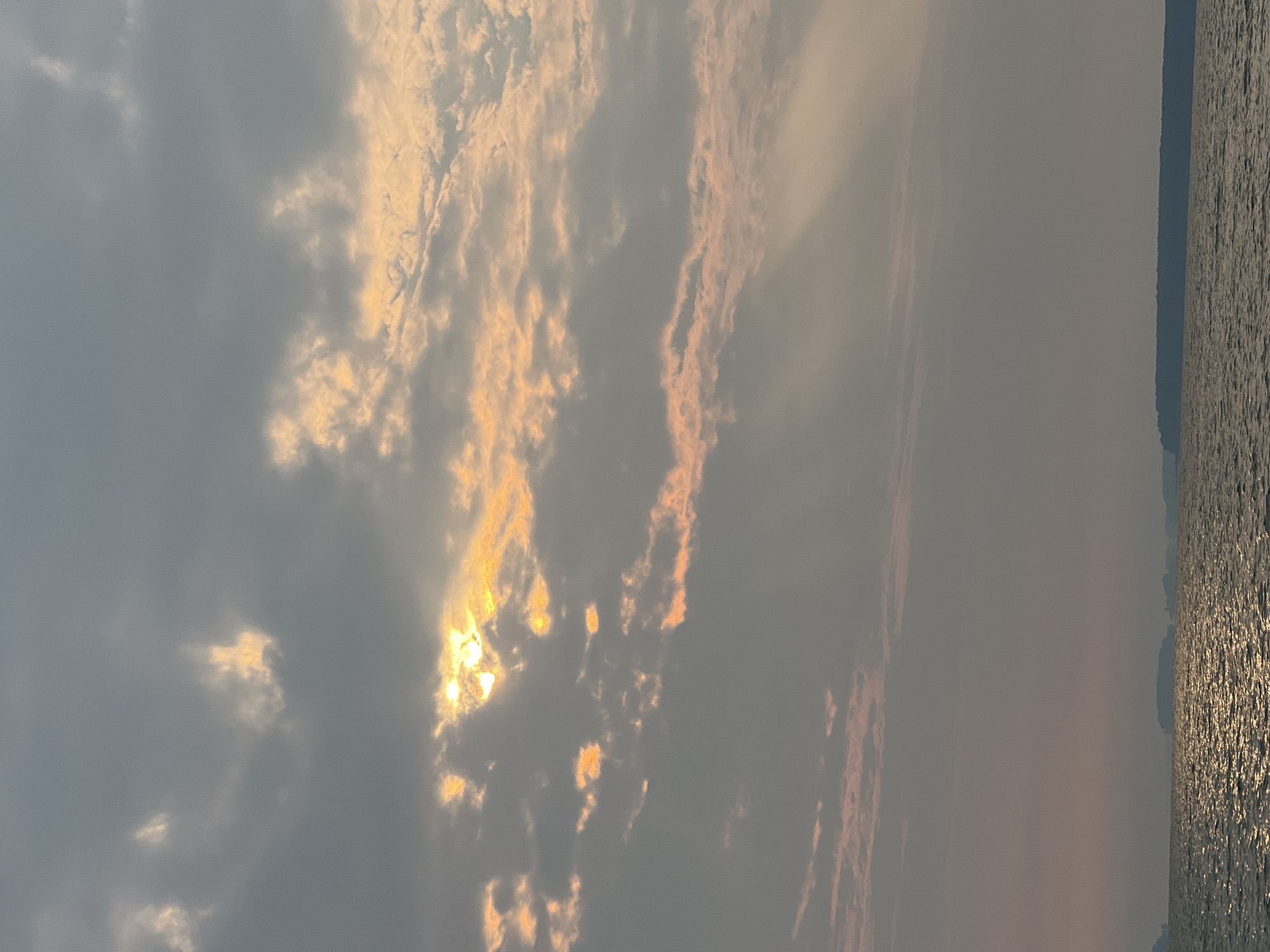 The image captures a body of water under a cloudy sky with the sun partially visible. The sun's rays are breaking through the clouds, casting an orange glow on the clouds and creating a shimmering effect on the water. The horizon shows distant landforms, appearing as dark silhouettes against the muted sky. The scene is likely a natural landscape, possibly a lake or sea, and the lighting suggests it could be during sunrise or sunset. The location is identified as Novopichugovo, Russia.