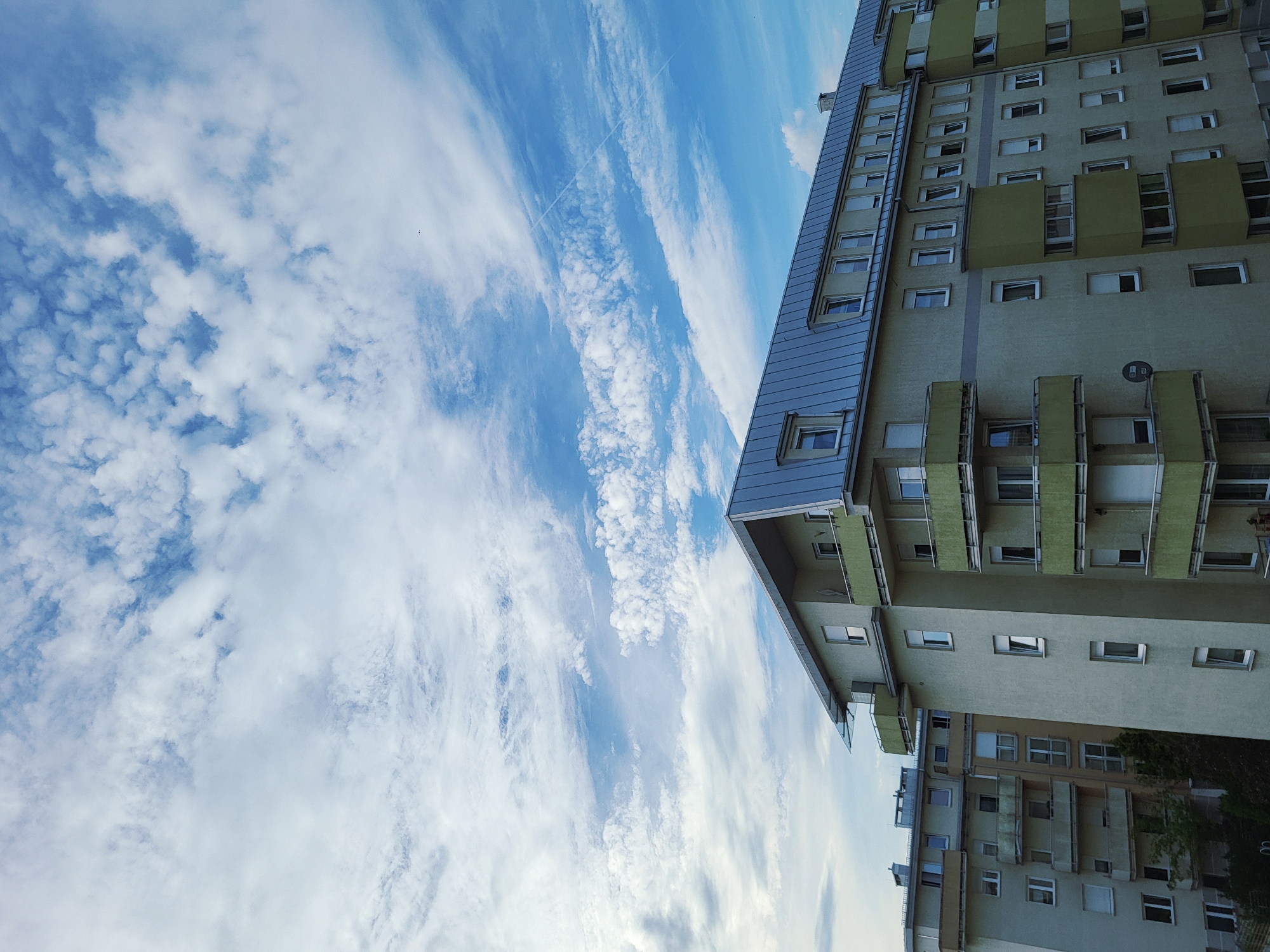 The image shows an exterior view of apartment buildings against a blue sky with scattered clouds. The buildings are modern in design, with light-colored facades and green balconies. One building in the foreground has a steeply pitched gray metal roof. There are no people or animals visible in the image. The overall impression is one of an urban residential area on a cloudy day.