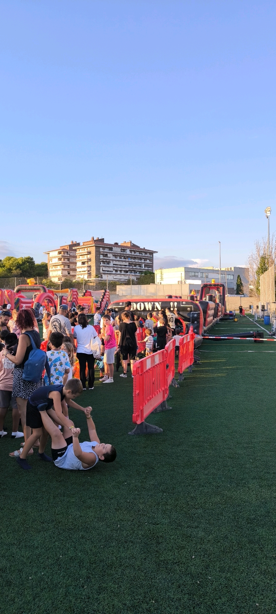 The image captures an outdoor event taking place on a green artificial turf field, likely a sports field, in Ripollet, Spain. The sky is clear and blue with a slight haze. In the background, a multi-story brick apartment building with balconies is visible, along with trees.
The foreground is occupied by a gathering of people, including children and adults, around inflatable attractions. There is a large, inflatable obstacle course that has the word "DOWN!!" written on it in large white letters on a black background. Red plastic barriers with white tape are set up to delineate areas or guide the crowd.
In the lower left quadrant, two young boys are interacting on the grass. One boy, wearing a light blue tank top and dark shorts, is lying on his back, with his legs in the air. Another boy, wearing a dark blue sleeveless shirt and shorts, is standing over him, holding his hands.
To the left of the inflatable obstacle course, a group of people are standing and observing. Several individuals are dressed in casual summer attire. A woman with red hair is wearing a black top and a patterned skirt, carrying a blue backpack. Other people are wearing t-shirts, shorts, and sandals.
In the middle ground, the inflatable obstacle course extends horizontally. People are queuing or moving around it. A bright red structure, likely part of another inflatable attraction, is visible to the left.
The overall atmosphere appears to be that of a community event or festival, possibly a summer fair or a children's party, set up in a public outdoor space within the city. The lighting suggests it is late afternoon or early evening.