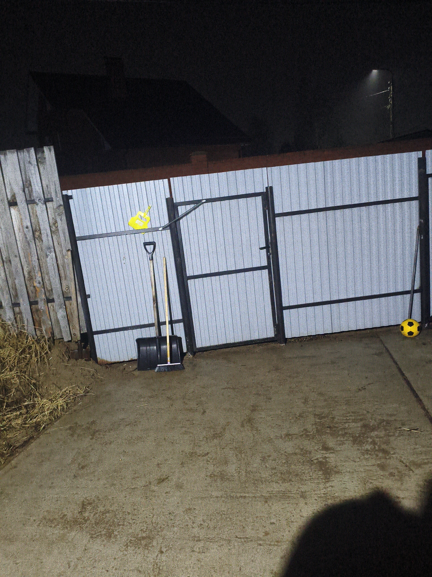 The image depicts an outdoor scene at night in Chaikovsky, Russia. A partially visible house with a dark roof is in the background. A metal fence runs across the middle of the frame, with a section made of corrugated metal and a gate. A wooden fence stands to the left of the metal fence. A yellow watering can is hanging on the metal fence, and a black snow shovel and broom lean against it. A yellow soccer ball rests near the right side of the fence. The ground is concrete. The lower right portion of the frame contains a dark shadow, suggesting a person or object obstructing the light source. A street lamp is visible in the upper right corner. Straw or dry grass is piled next to the wooden fence.